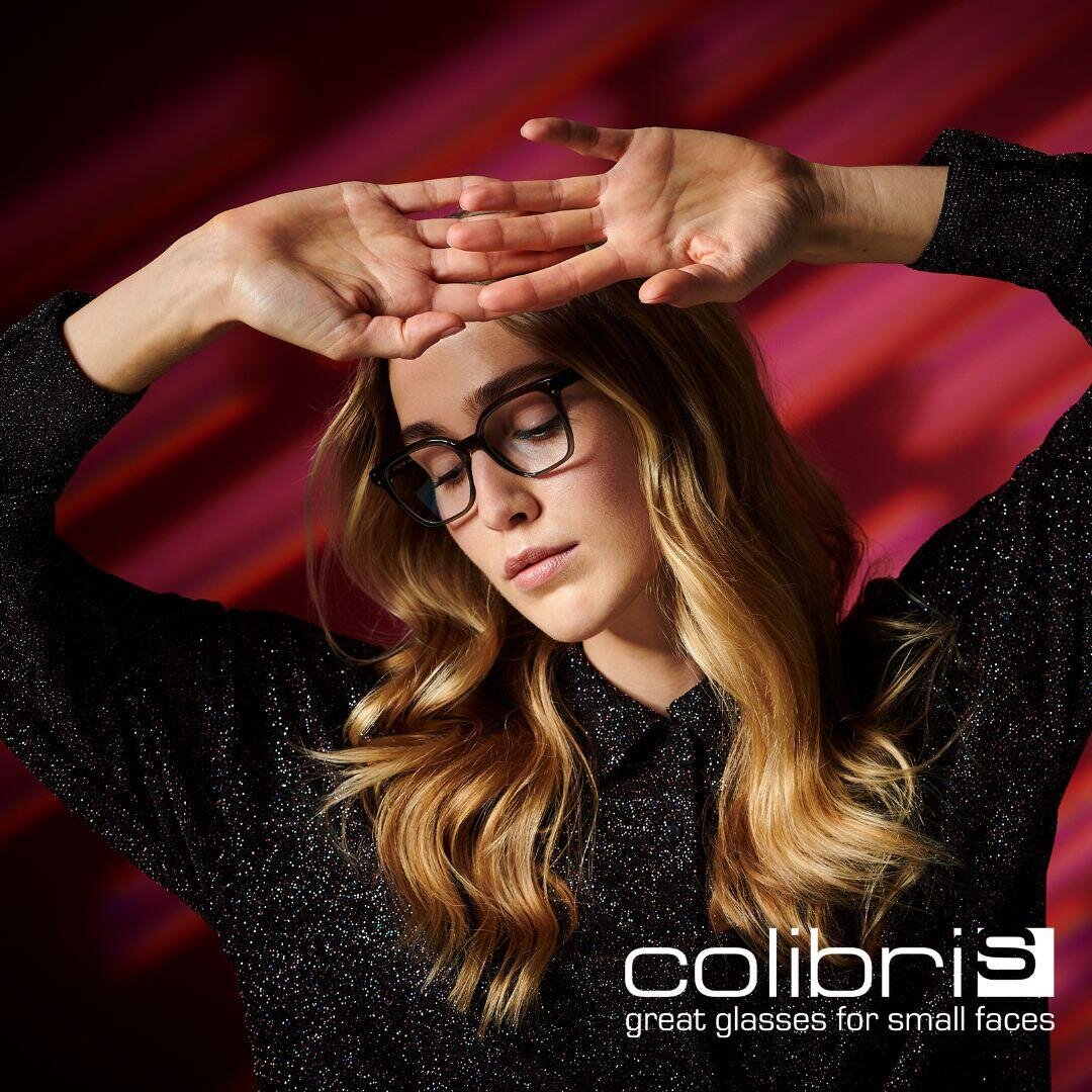 A woman with long blonde hair wearing black glasses poses with her hands above her head in front of a red and pink abstract background. The text reads: colibris – great glasses for small faces.