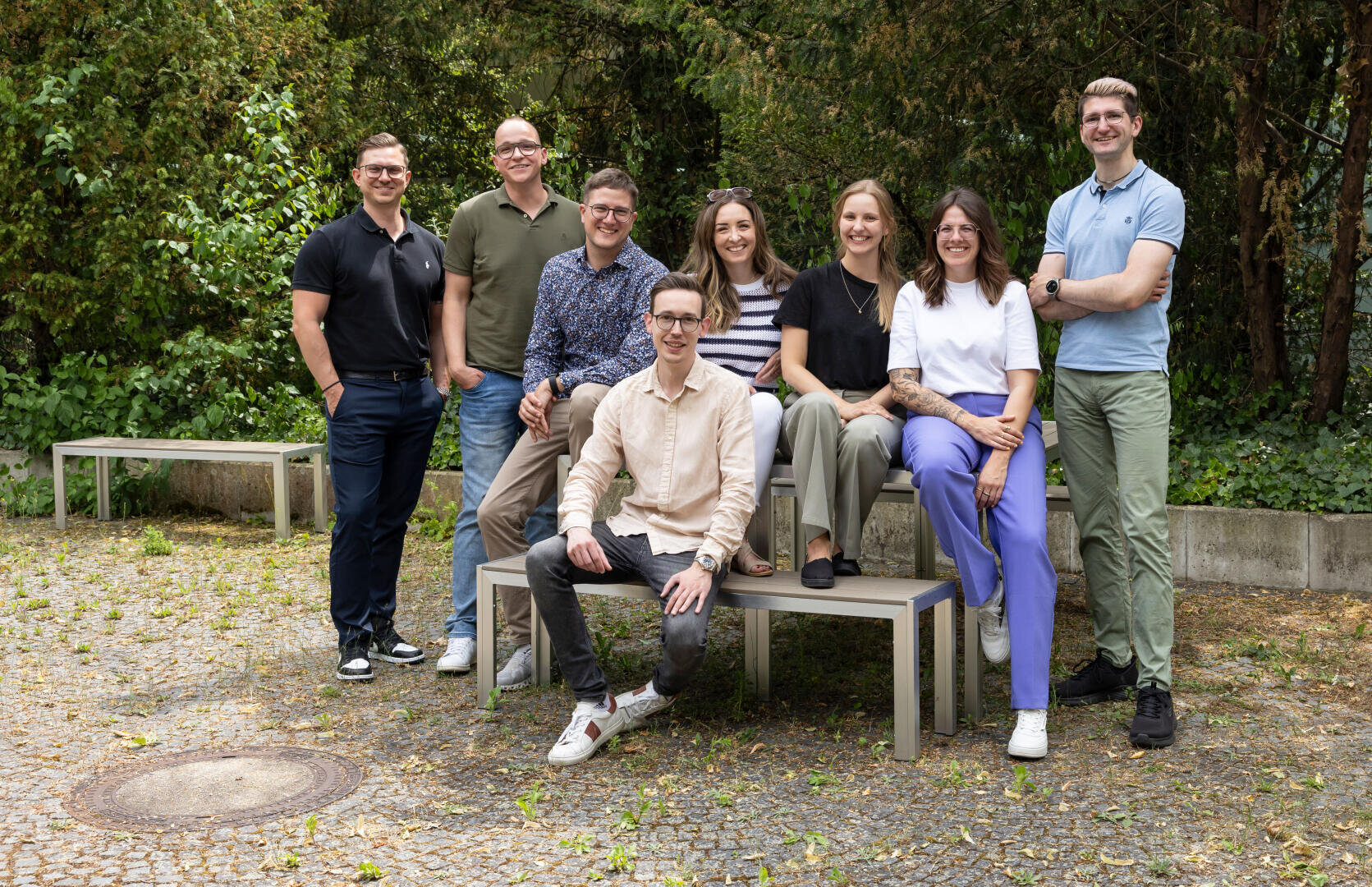 Eight people pose and smile outdoors in casual clothes; five stand behind them and three sit on benches. Trees and green plants form a natural background. The atmosphere is friendly and relaxed.