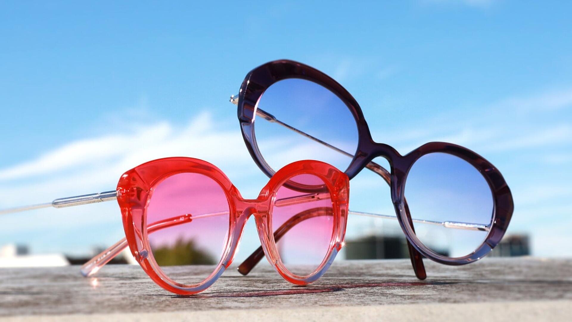 Two pairs of round sunglasses, one with pink frames and lenses and the other with purple frames and blue-tinted lenses, rest on a wooden surface under a bright blue sky.
