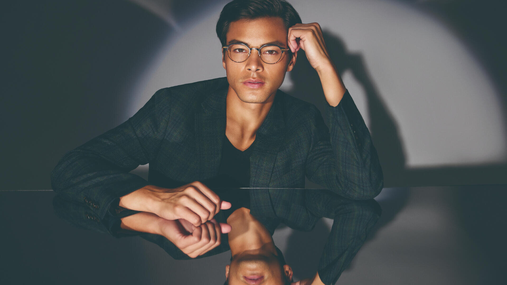 Ein Mann mit Brille und dunklem Blazer sitzt an einem spiegelnden Tisch und blickt direkt in die Kamera. Seine Hand ruht auf seiner Stirn. Sein Spiegelbild ist auf der glänzenden Oberfläche sichtbar.