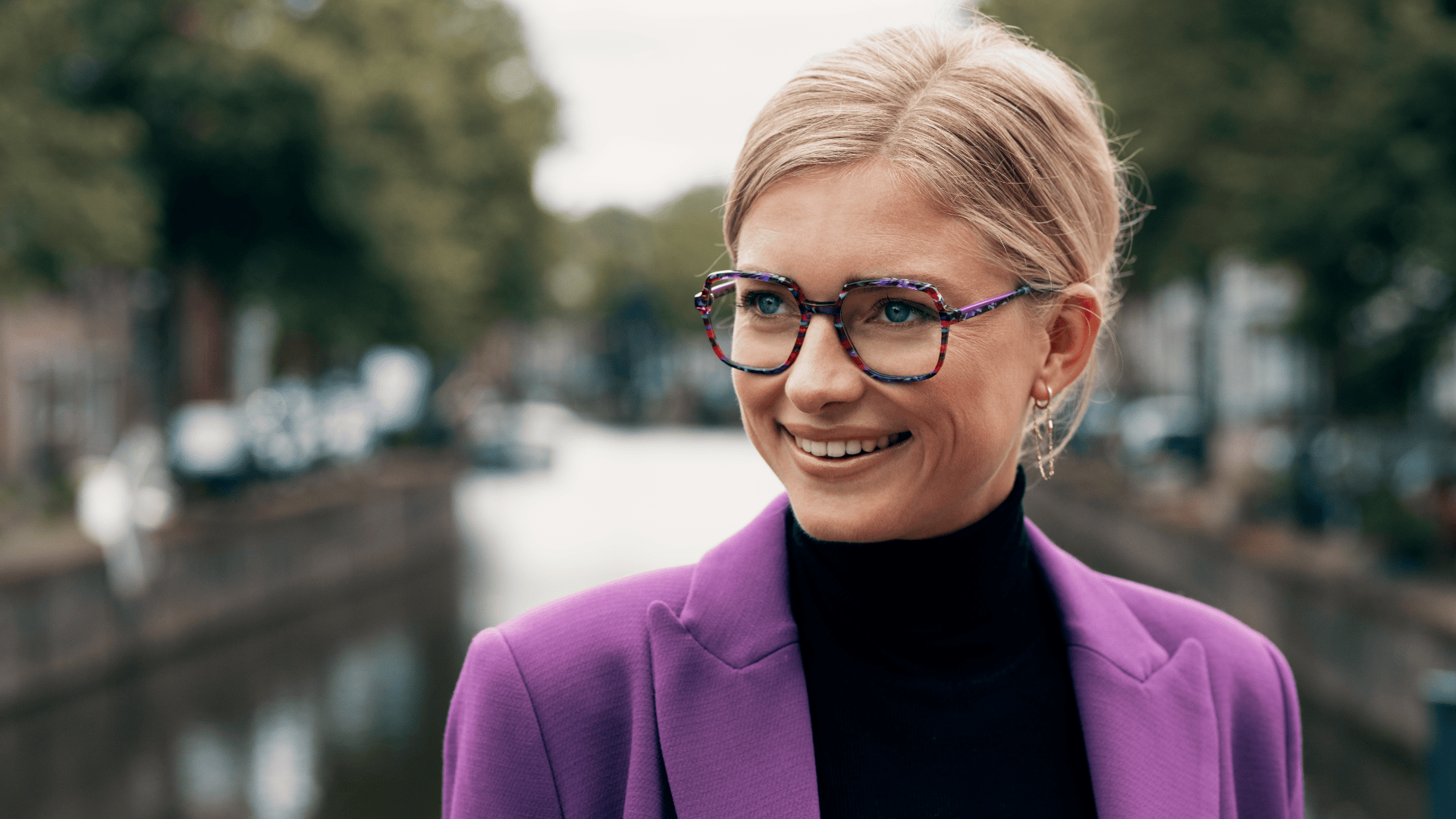 Eine lächelnde Frau mit kurzen blonden Haaren, die eine bunte Brille, einen schwarzen Rollkragenpullover und einen lila Blazer trägt, steht im Freien in der Nähe eines Kanals mit Bäumen und Gebäuden im Hintergrund.
