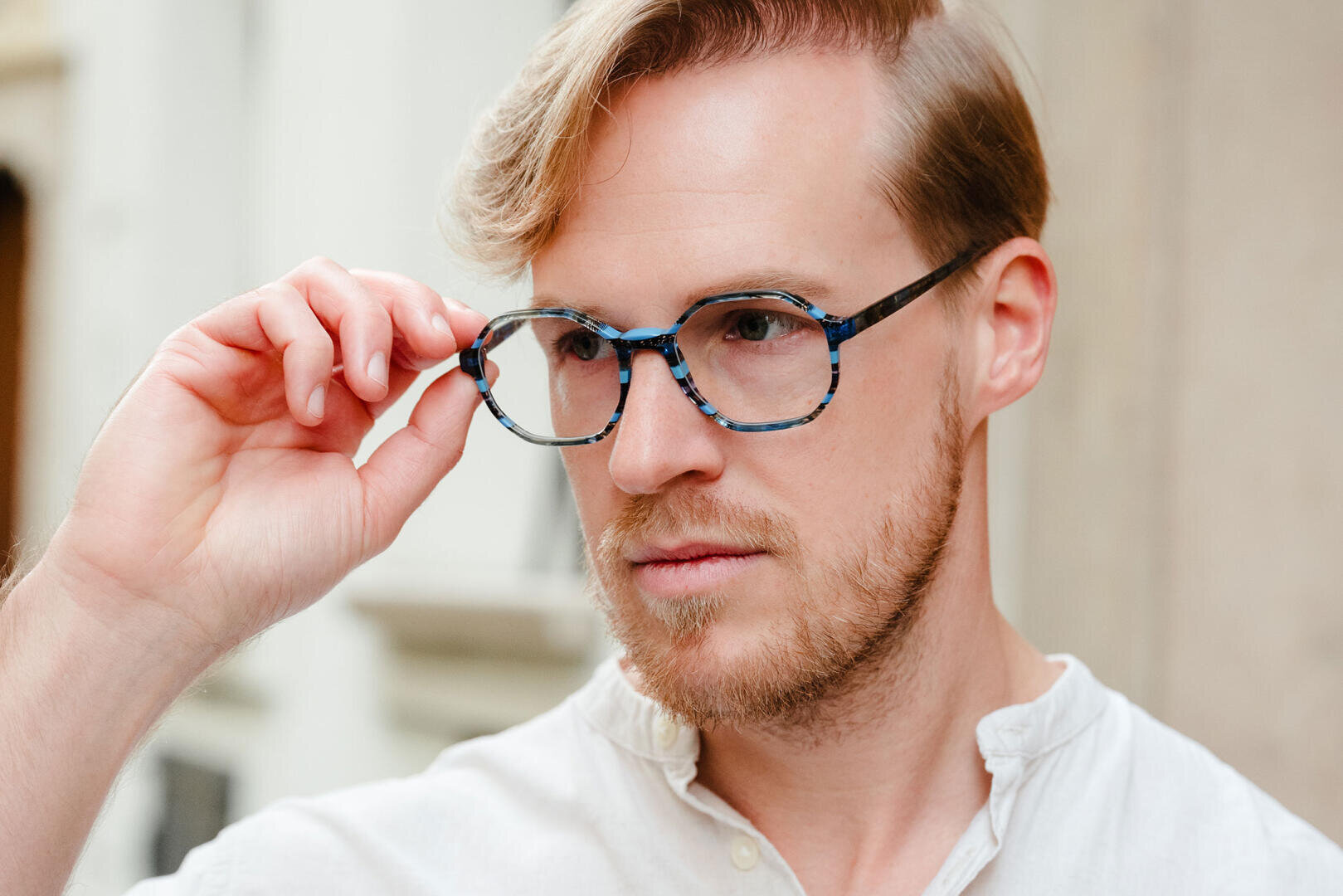 Ein Mann mit hellbraunem Haar, kurzem Bart und einer Brille mit blauem Schildpattrahmen rückt seine Brille zurecht, während er nach vorne blickt. Er trägt ein weißes Hemd und befindet sich im Freien in einer leicht verschwommenen städtischen Umgebung.