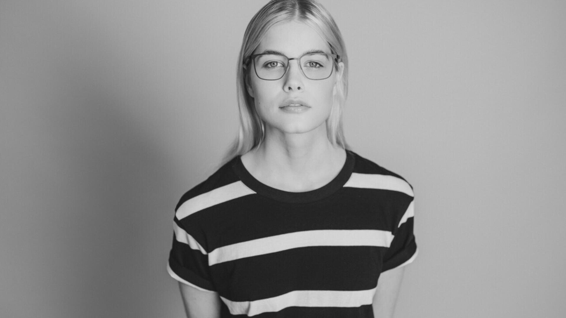Black-and-white photo of a person with long blond hair and glasses, wearing a black T-shirt with white horizontal stripes, standing against a plain background and looking directly at the camera.