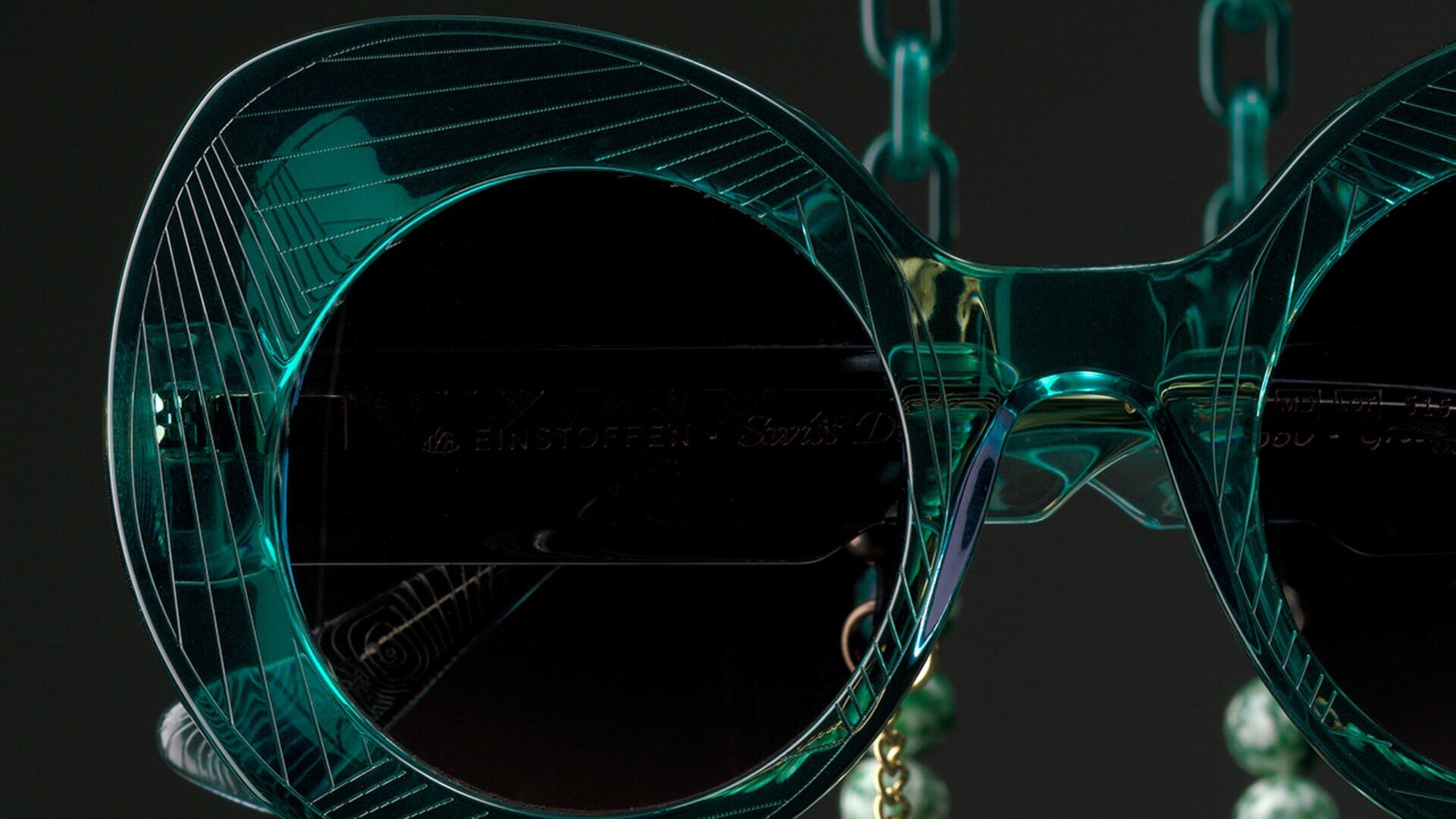Nahaufnahme einer stilvollen, runden, blaugrünen Sonnenbrille mit dunklen Gläsern und geometrischen Mustern auf dem Rahmen vor dunklem Hintergrund. An der Brille ist eine grüne Kette befestigt.