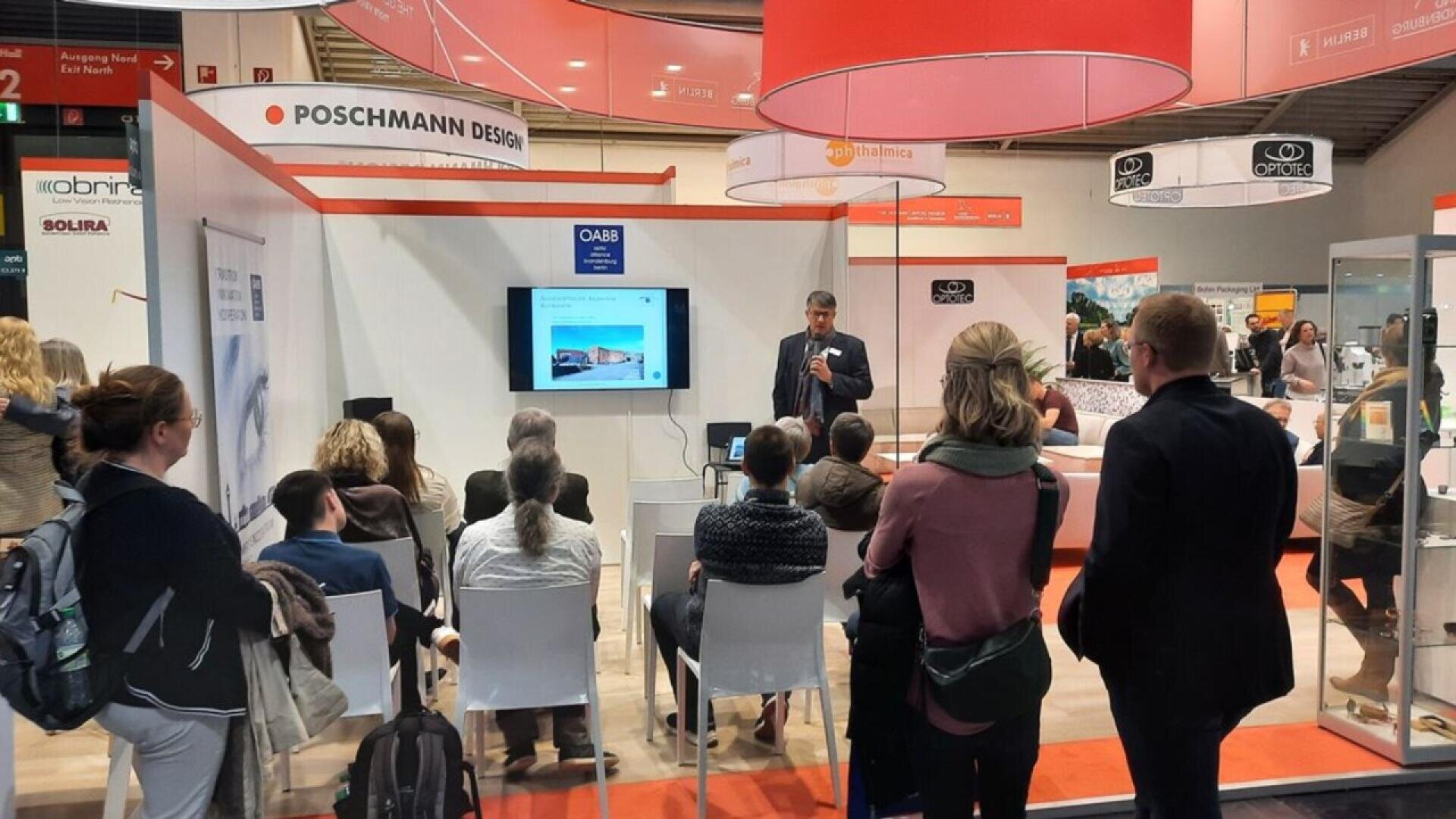 A man gives a presentation to a seated audience at a trade show booth with banners and display stands. People listen attentively, and a screen shows a slide with an image and text. The booth is labeled POSCHMANN DESIGN.