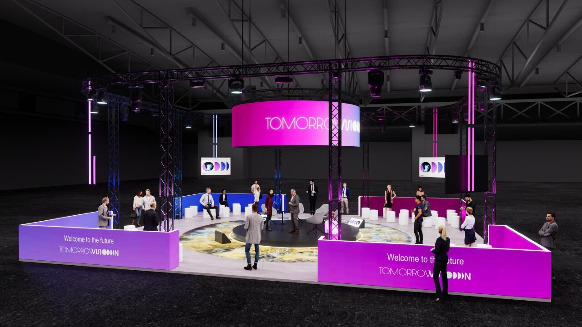 A modern event setup features people networking and conversing around a circular stage with digital screens displaying “TOMORROW.” The area is lit with purple and pink lights under a large metal truss in a spacious hall.