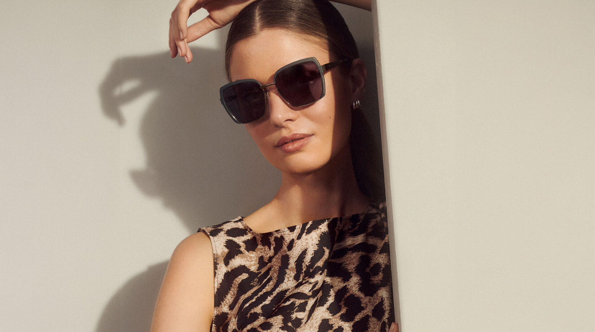 Eine Frau mit einer großen schwarzen Sonnenbrille und einem ärmellosen Oberteil mit Leopardenmuster lehnt an einer hellen Wand, die Haare nach hinten gezogen und eine Hand über dem Kopf.