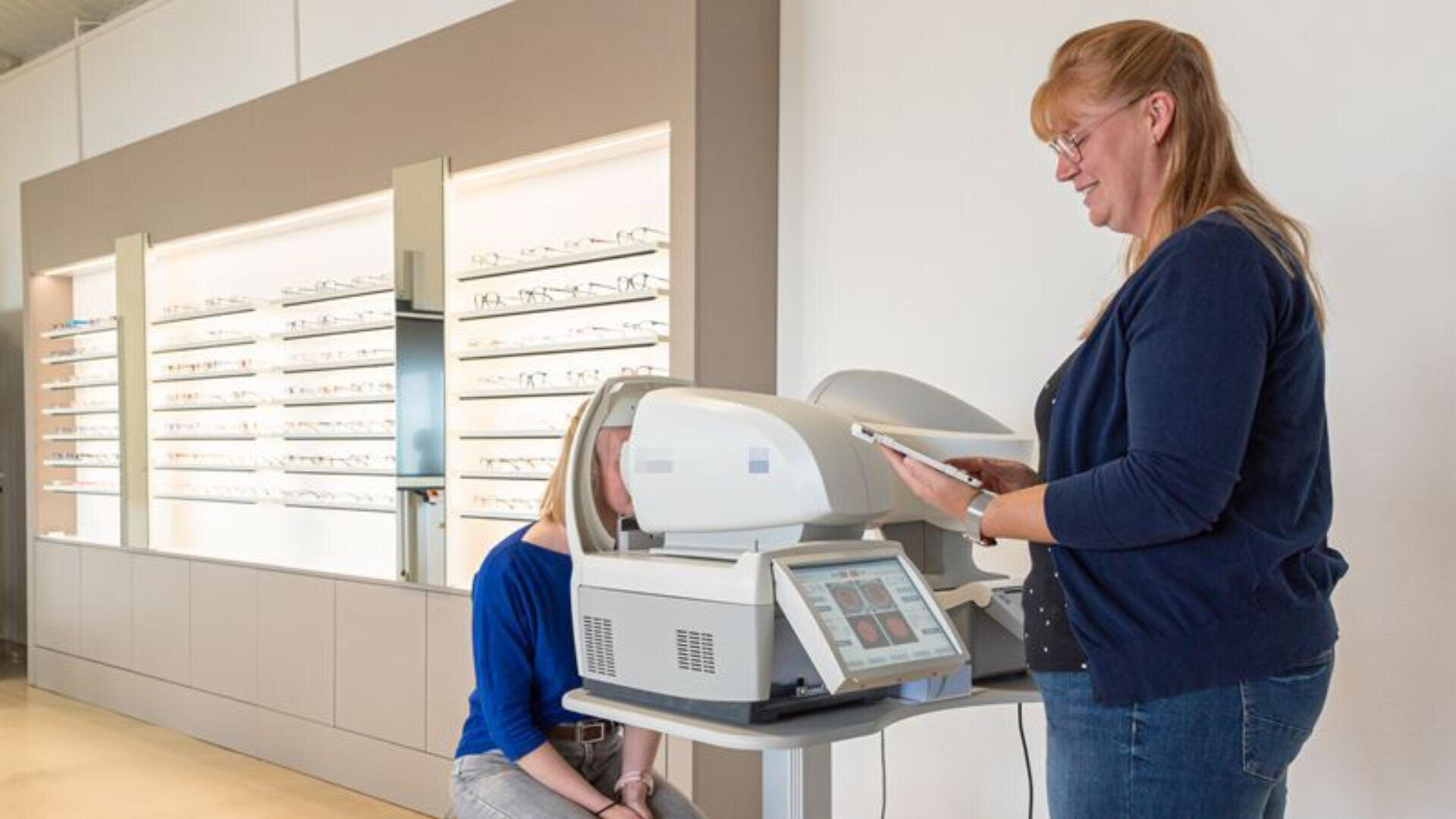 Eine Frau bedient in der Praxis eines Optikers ein Gerät zur Augenuntersuchung an einem sitzenden Patienten. Im Hintergrund sind Regale mit Brillen ausgestellt.