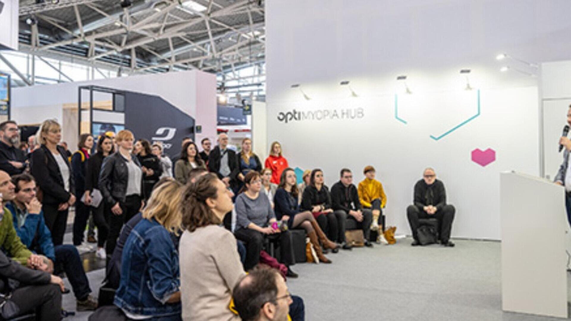 A group of people sits and stands attentively in a modern, well-lit exhibition space, listening to a speaker at a podium near a white wall with OPTI MORAHUB and a small pink heart symbol.