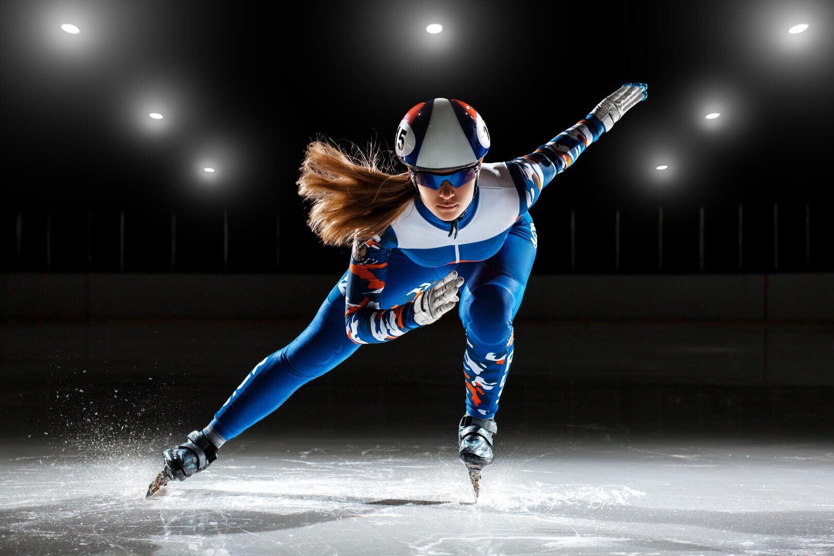 Ein Eisschnellläufer in blau-weißem Anzug, Helm und Brille gleitet in einer dynamischen Action-Pose auf einer Eislaufbahn unter hellem Licht.
