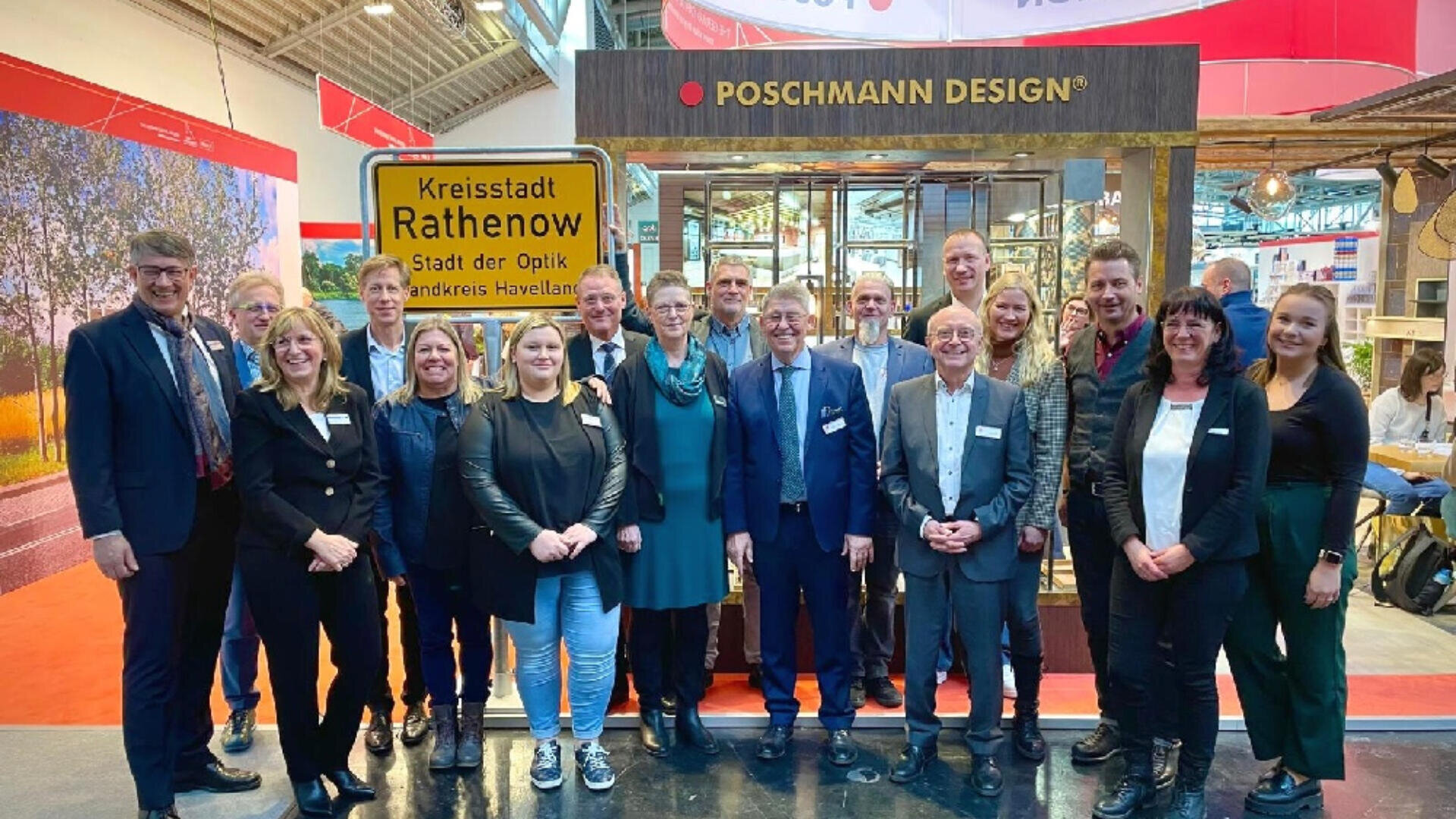 Eine Gruppe von Menschen steht lächelnd vor einem Stand mit einem „Kreisstadt Rathenow“-Schild und einem Banner von Poschmann Design, scheinbar auf einer Indoor-Ausstellung oder Messe.