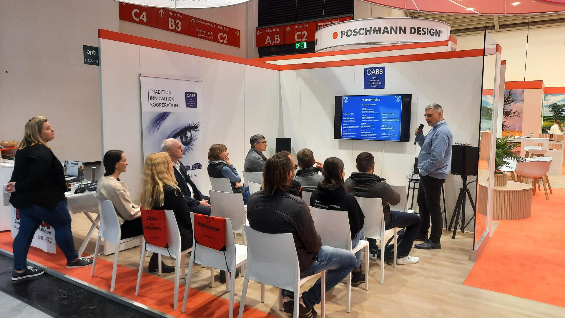 A group of people sit on white chairs listening to a man giving a presentation in a modern exhibition booth labeled POSCHMANN DESIGN. A large screen displays information, and banners are visible on the white walls.
