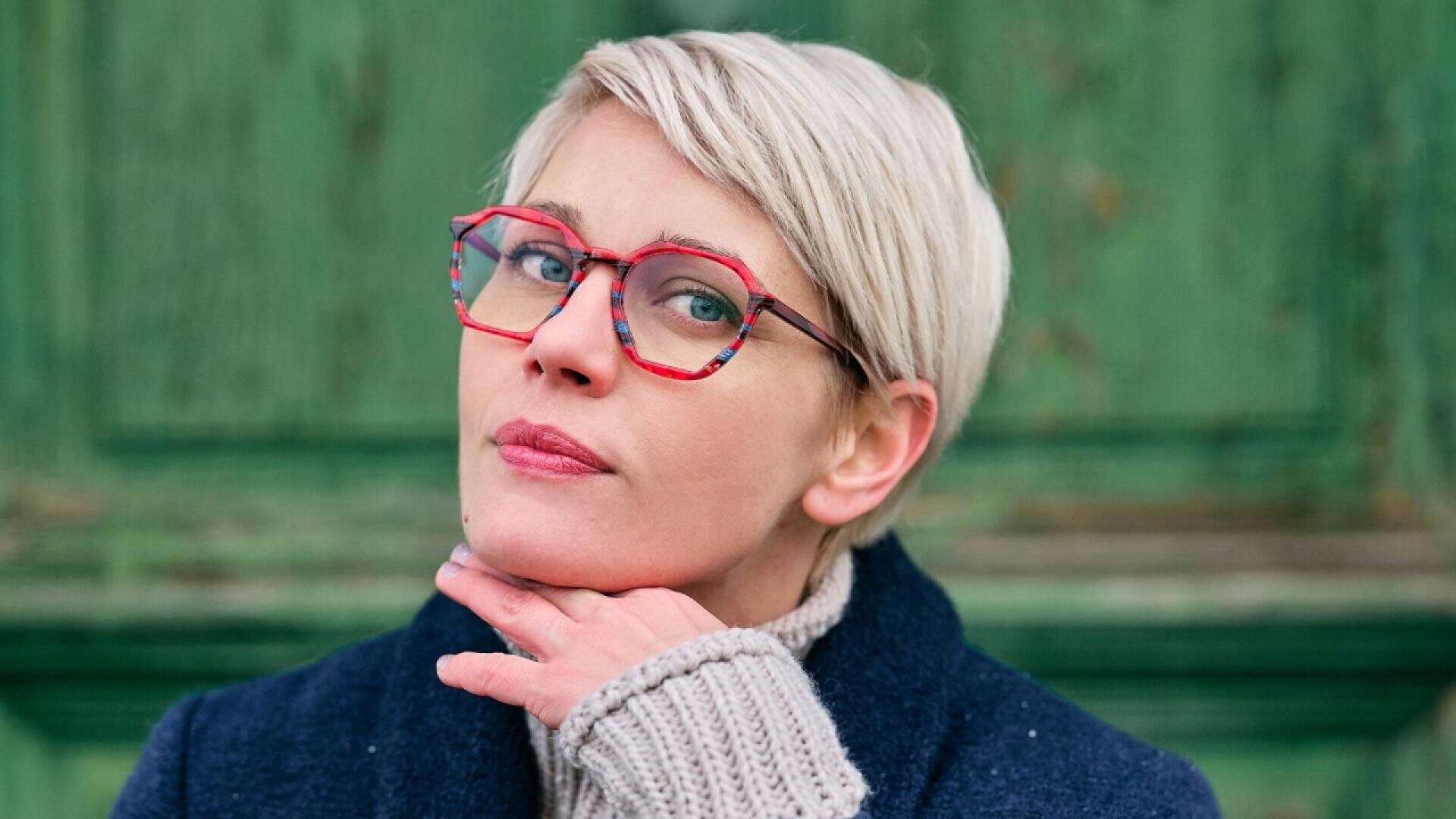 A woman with short blonde hair and colorful red glasses poses with her chin resting on her hand. She is wearing a navy coat and a gray knit sweater against a green wooden background.