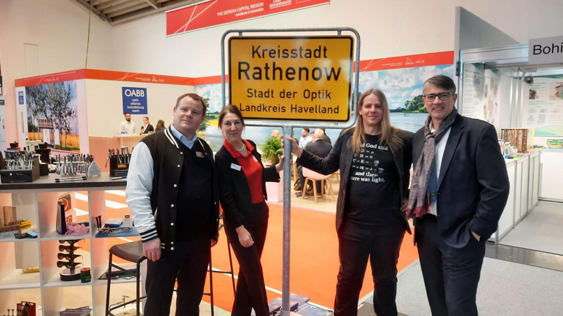 Vier Personen stehen lächelnd im Innenbereich einer Veranstaltung und posieren um ein gelbes Ortsschild mit der Aufschrift „Kreisstadt Rathenow, Stadt der Optik, Landkreis Havelland“. Im Hintergrund sind Messestände und Displays zu sehen.