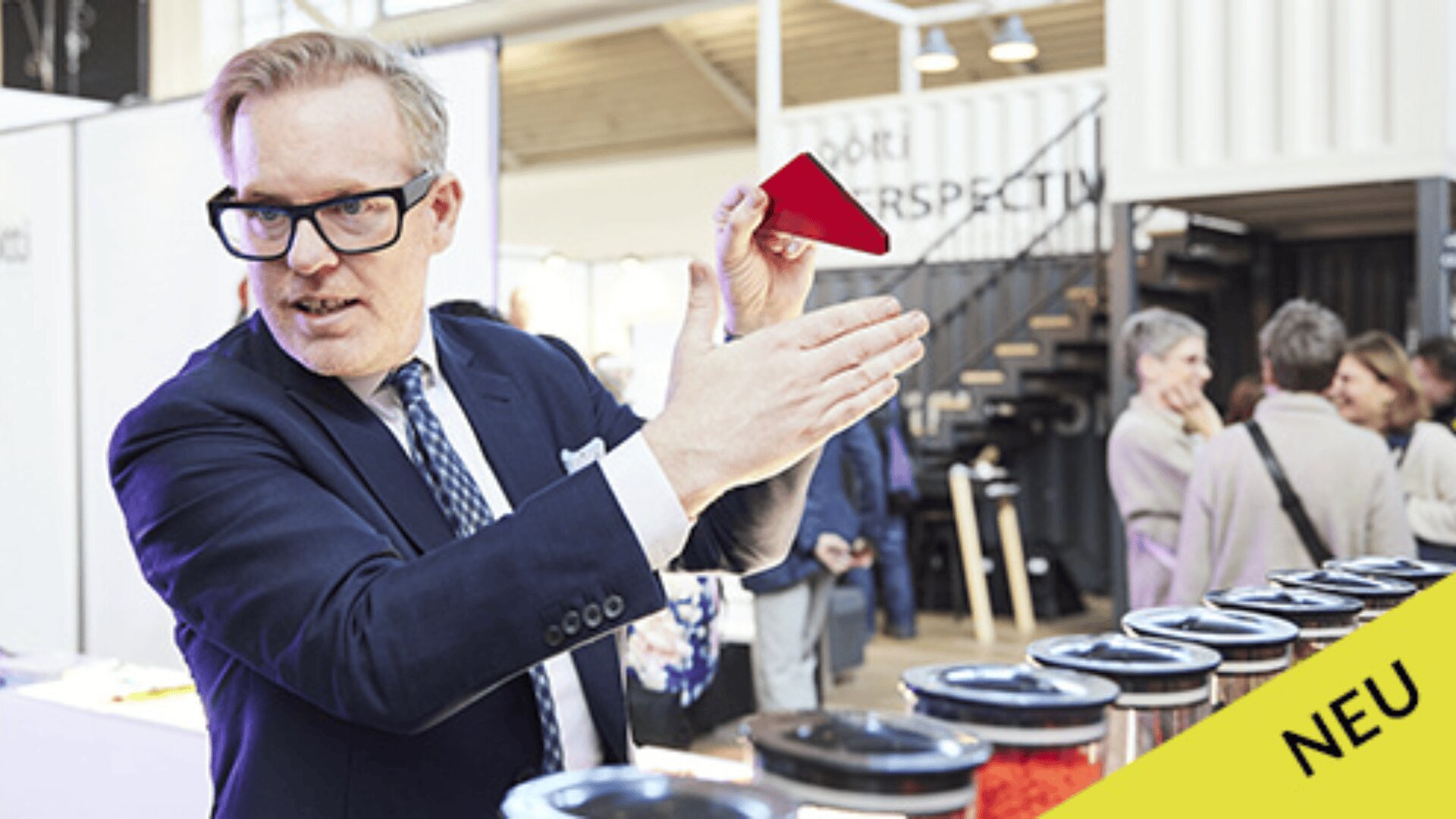 Ein Mann im Anzug gestikuliert während einer Indoor-Veranstaltung neben einer Reihe von Containern. Im Hintergrund sind Menschen und Exponate zu sehen. In der Ecke steht auf einem gelben Etikett „NEU“.