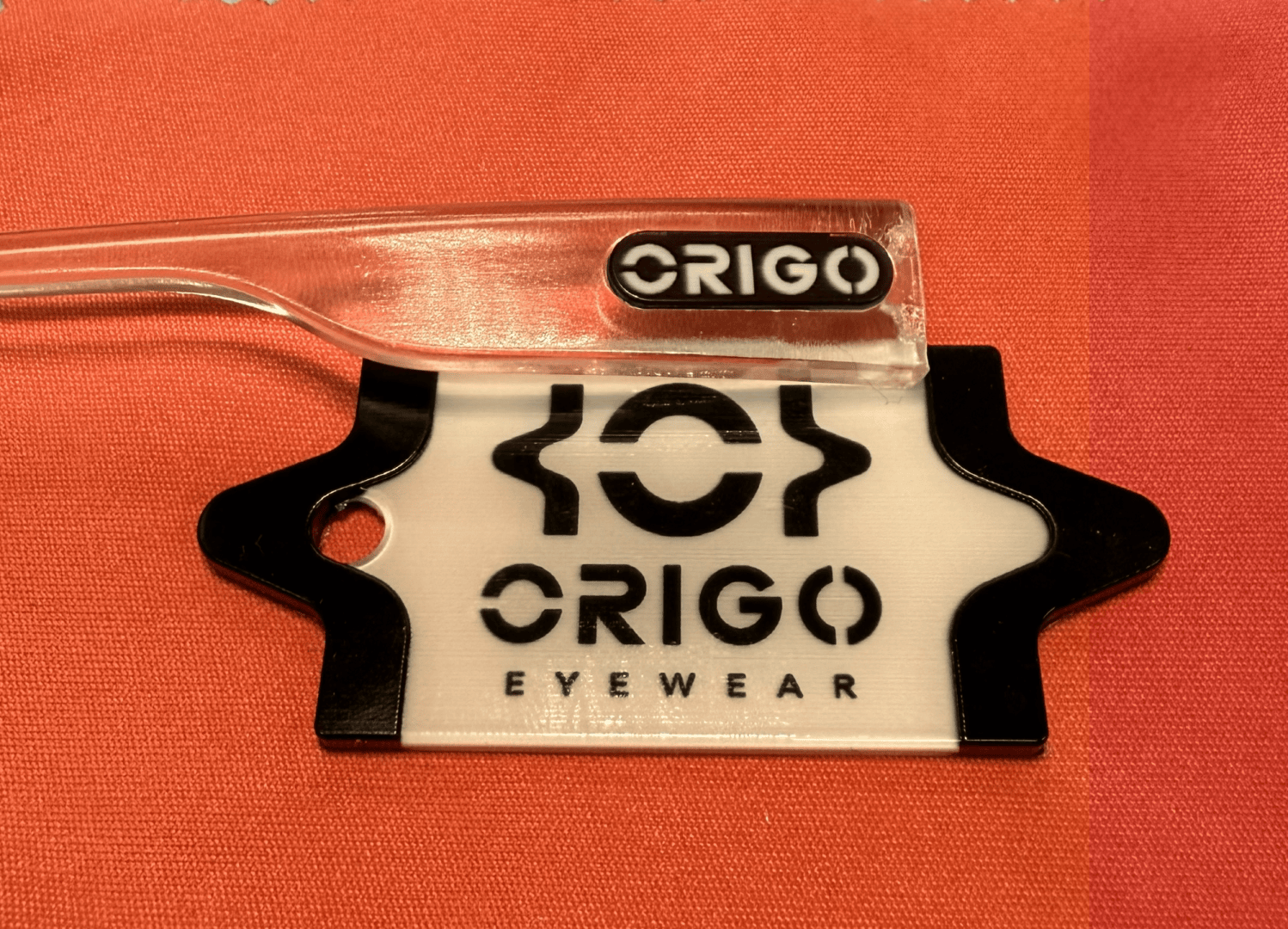 A transparent frame with the ORIGO logo rests on a white and black display card of the ORIGO EYEWEAR brand, which is placed on a red and orange fabric surface.