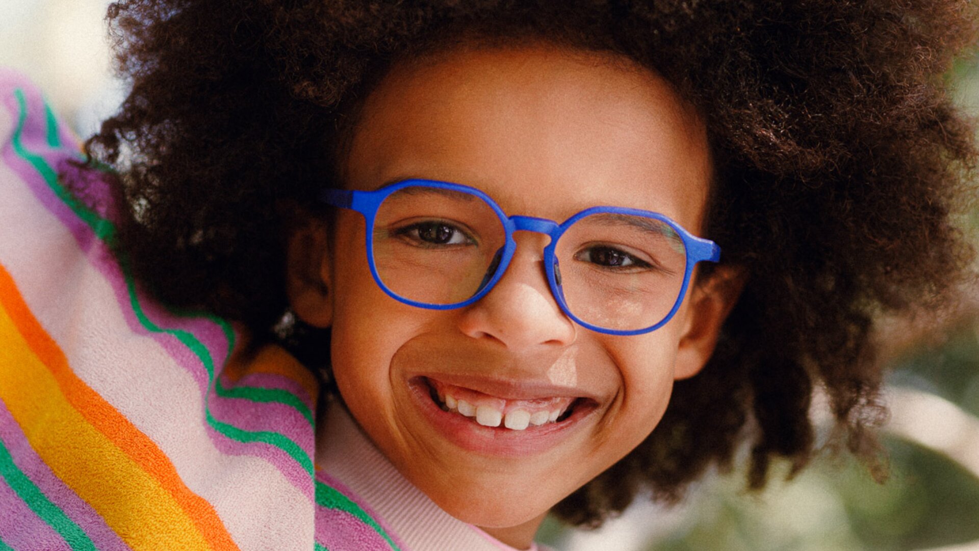 Lächelndes Kind mit lockigem Haar, das eine hellblaue Brille und einen bunt gestreiften Pullover trägt und direkt in die Kamera schaut.