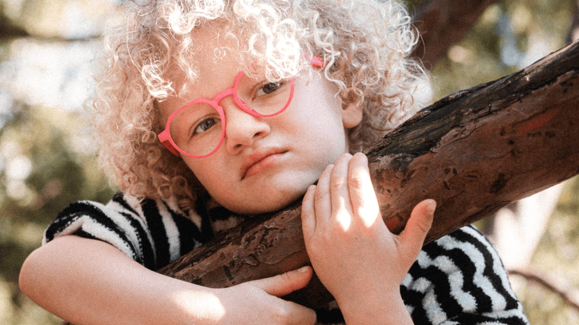 Ein kleines Kind mit lockigem blondem Haar und rosa Brille lehnt nachdenklich an einem Ast. Das Kind trägt ein schwarz-weiß gestreiftes Hemd, und das Sonnenlicht fällt durch die Bäume im Hintergrund.