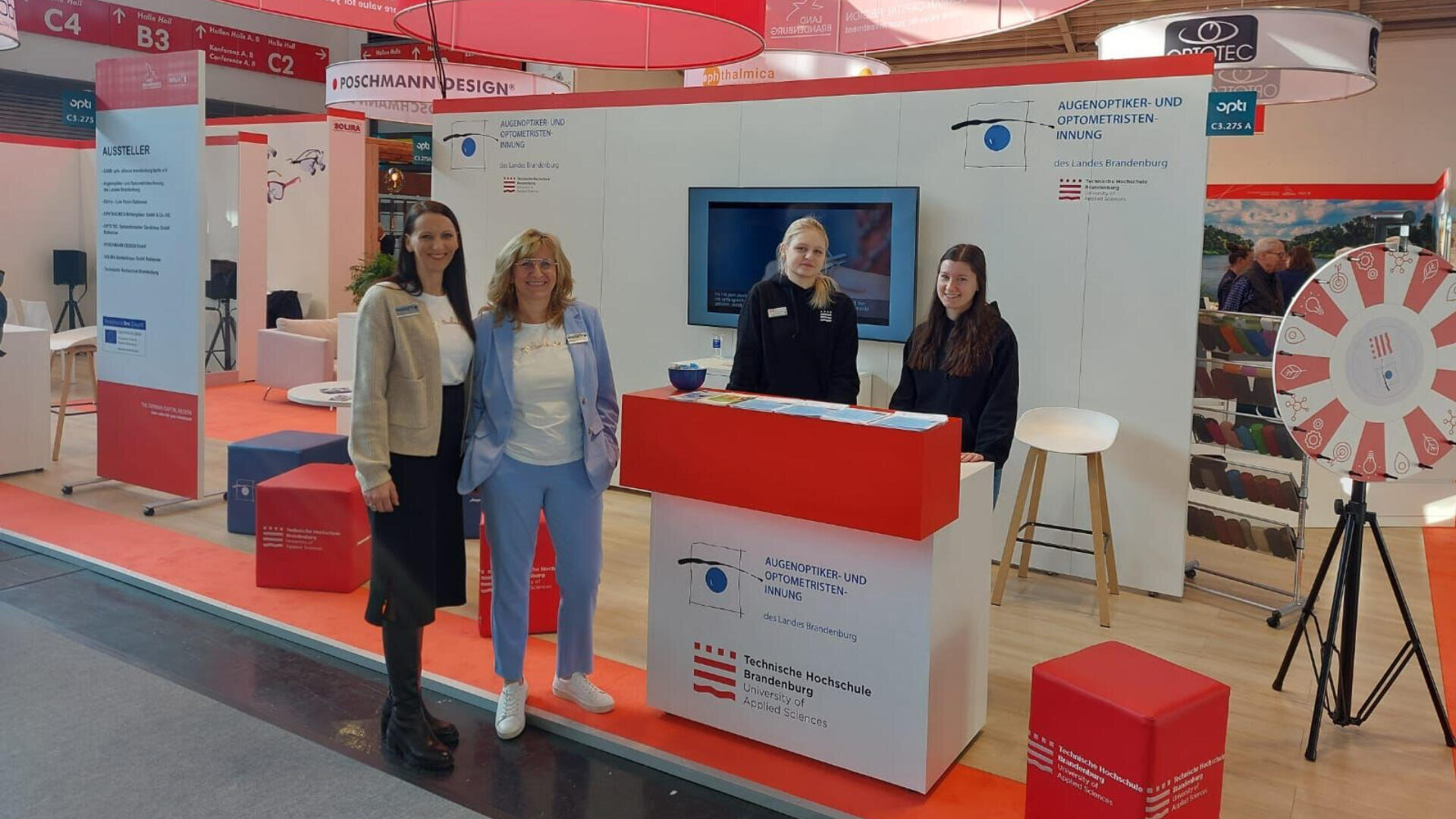 Vier Frauen stehen lächelnd an einem Messestand mit Informationsdisplays, einer roten Theke, einem sich drehenden Glücksrad und Bannern in einem hellen Veranstaltungsraum mit rot-weißem Dekor.