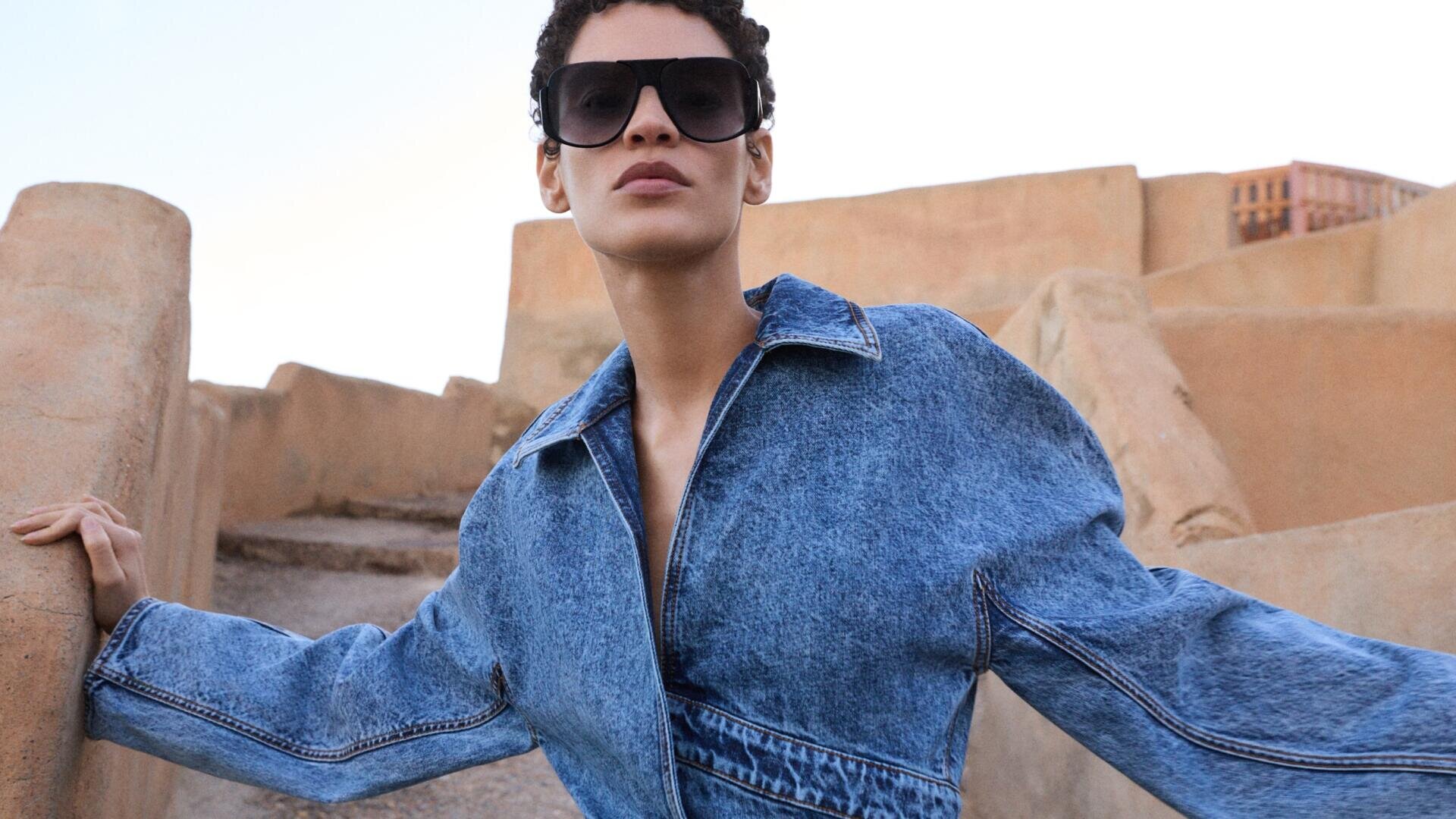 Eine Person, die eine große Sonnenbrille und eine blaue Jeansjacke trägt, steht im Freien vor einem Hintergrund aus hellbraunen Steinstufen und -wänden und blickt selbstbewusst in die Kamera.