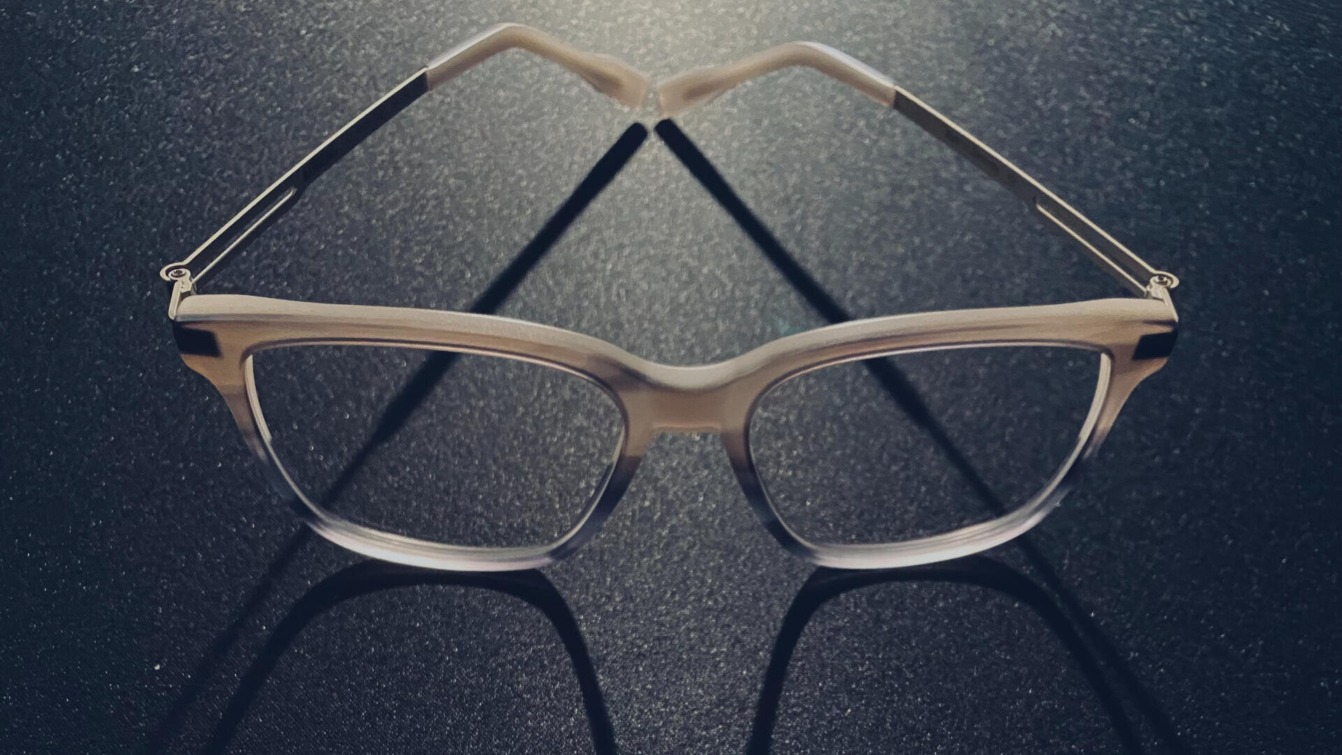 Eine Brille mit klarem Rahmen liegt auf einer dunklen, strukturierten Oberfläche und wirft deutliche Schatten, die eine herzähnliche Form bilden.