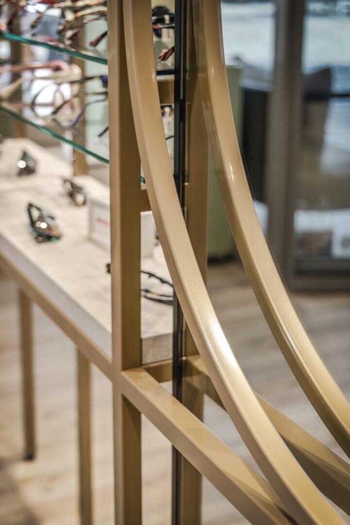 Nahaufnahme eines modernen Brillenständers mit goldenen Metallbögen und Glasböden, auf denen verschiedene Brillen in einem hell erleuchteten Ladeninneren aufbewahrt werden.