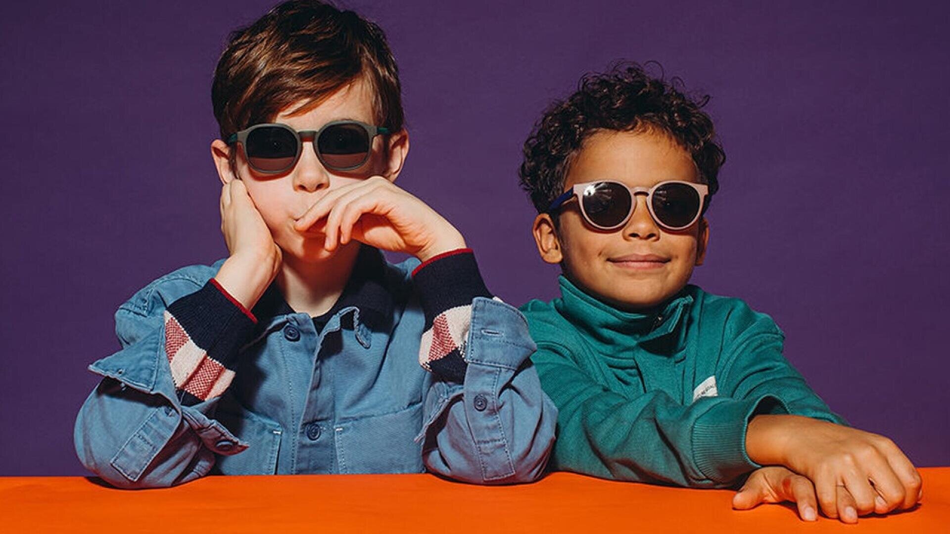 Two young boys sit side by side at an orange table, both wearing sunglasses. One boy rests his chin on his hand, while the other smiles slightly. They are set against a purple background.