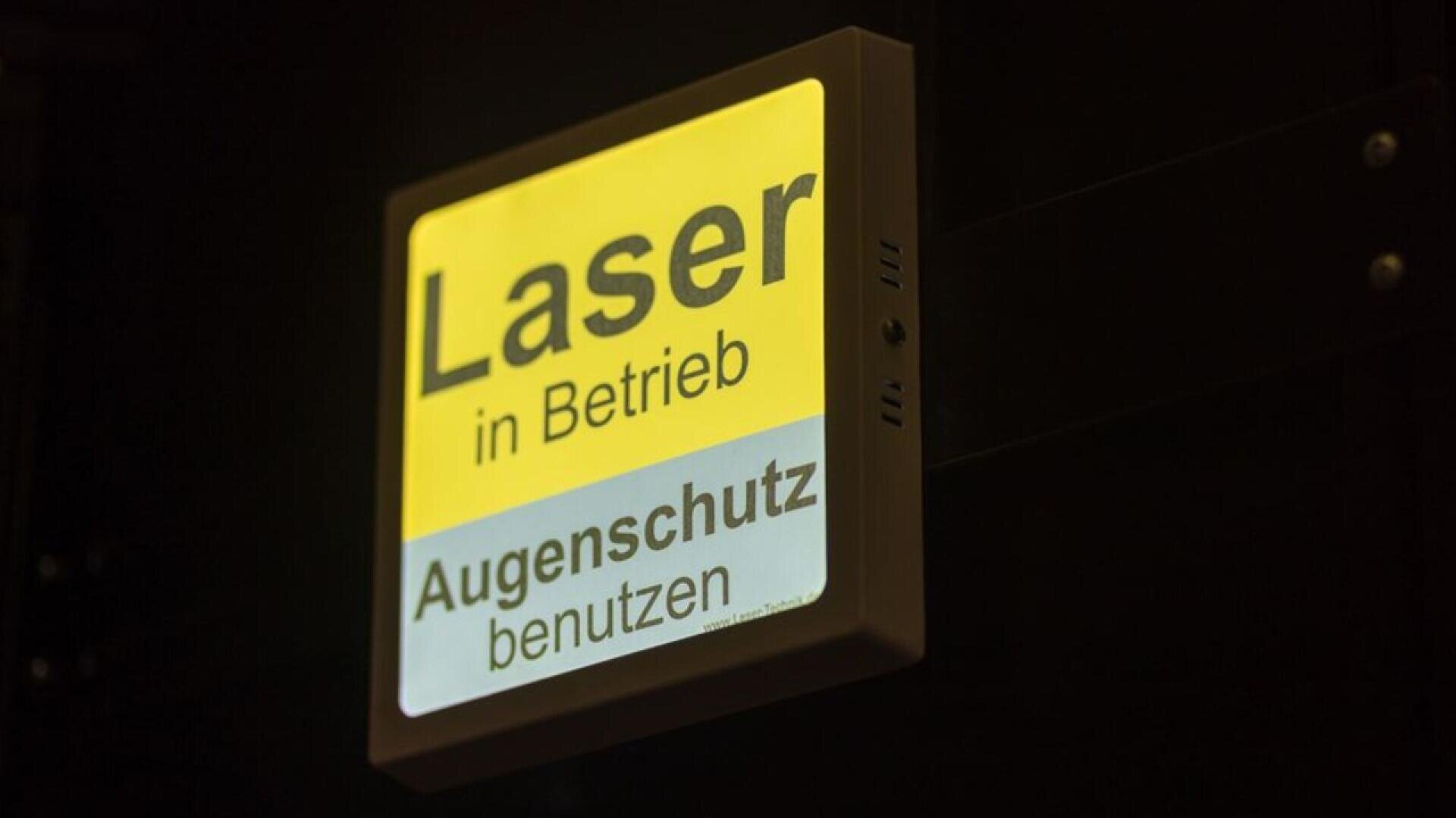 A square sign with yellow and white background reads Laser in Betrieb and Augenschutz benutzen, warning in German that a laser is in operation and eye protection must be used.