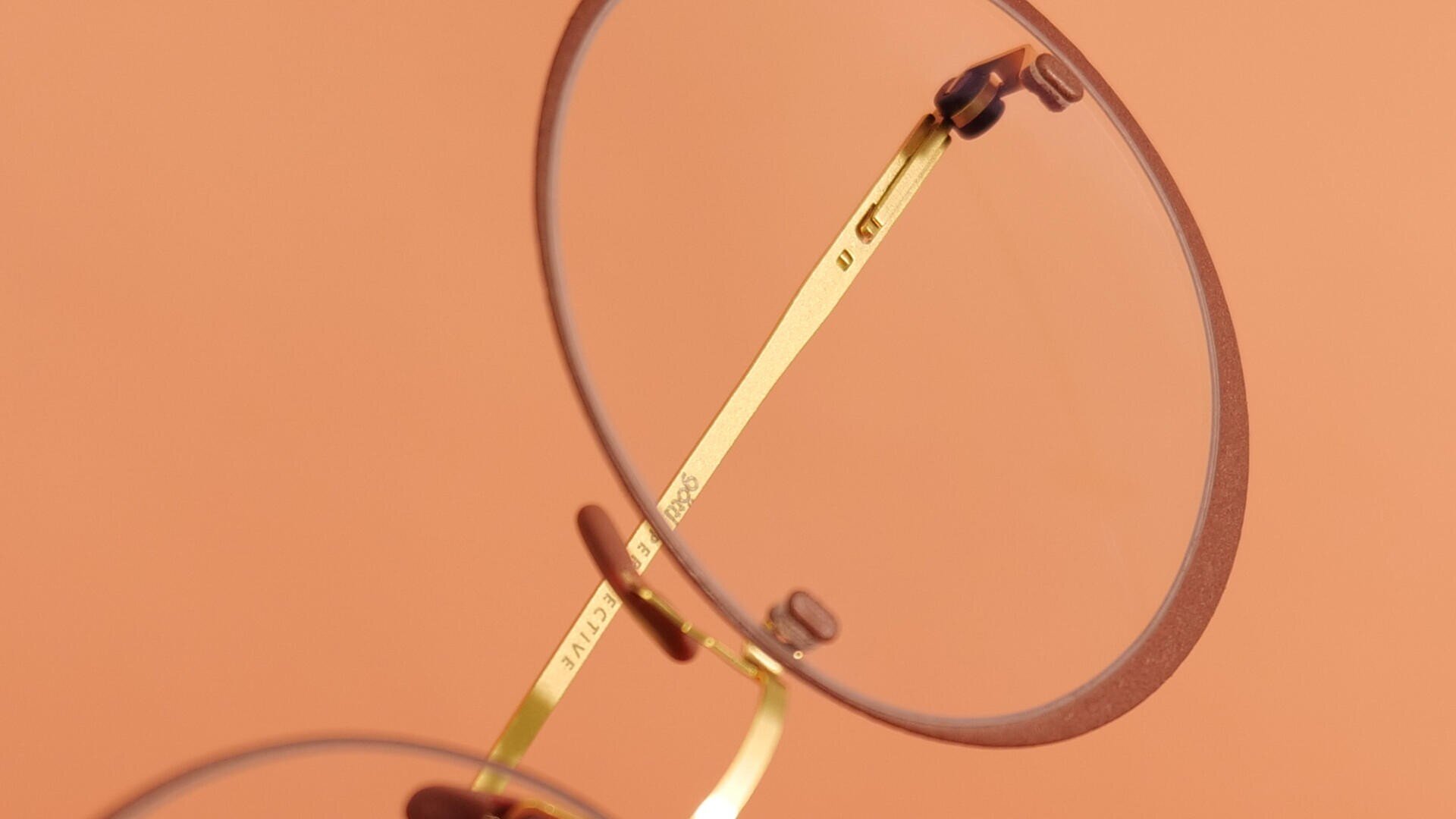 Nahaufnahme einer runden Brille mit dünnem Goldrahmen und klaren Gläsern vor einem zartpfirsichfarbenen Hintergrund. Der Fokus liegt auf dem Steg und den Nasenpads der Brille.