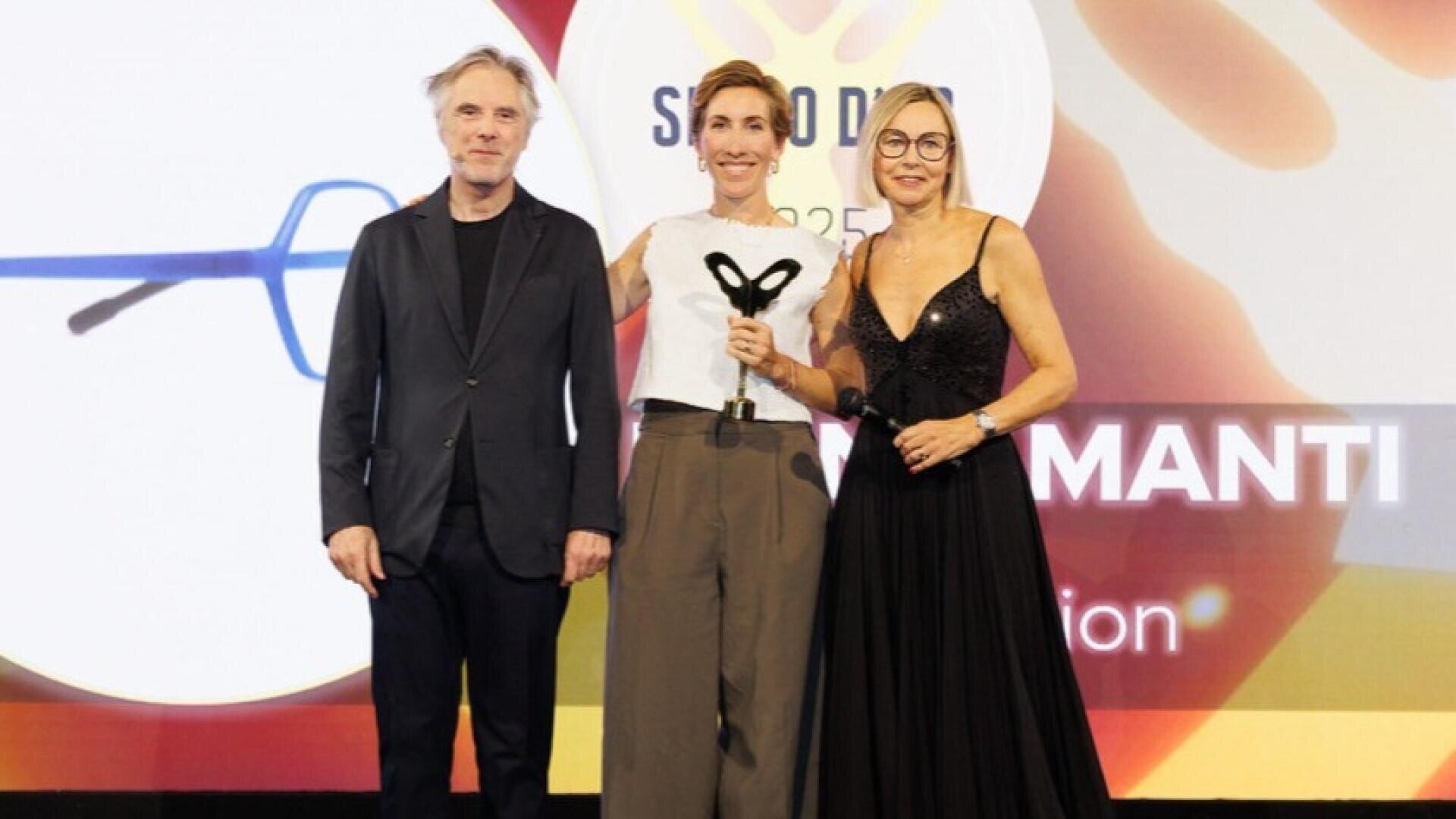 Three people stand on stage at an awards ceremony. The woman in the middle is holding a trophy. A colorful screen with text and graphics can be seen in the background. All three people are smiling and formally dressed.