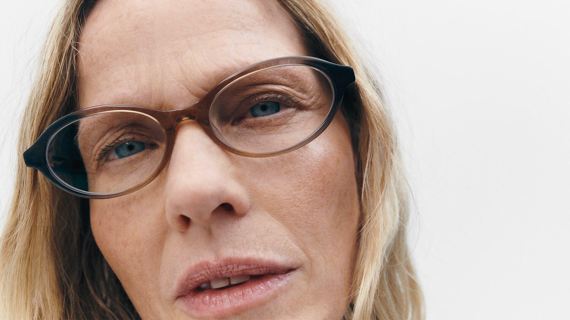 Close-up of the face of a person with fair skin, blue eyes, and blond hair, wearing brown-rimmed glasses and looking directly at the camera against a plain white background.