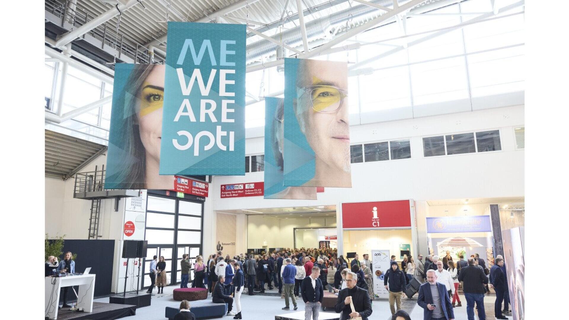 A large indoor event with many people walking and gathering. Three hanging banners show faces and text reading ME WE ARE opti. The venue is bright with high ceilings and modern architecture.