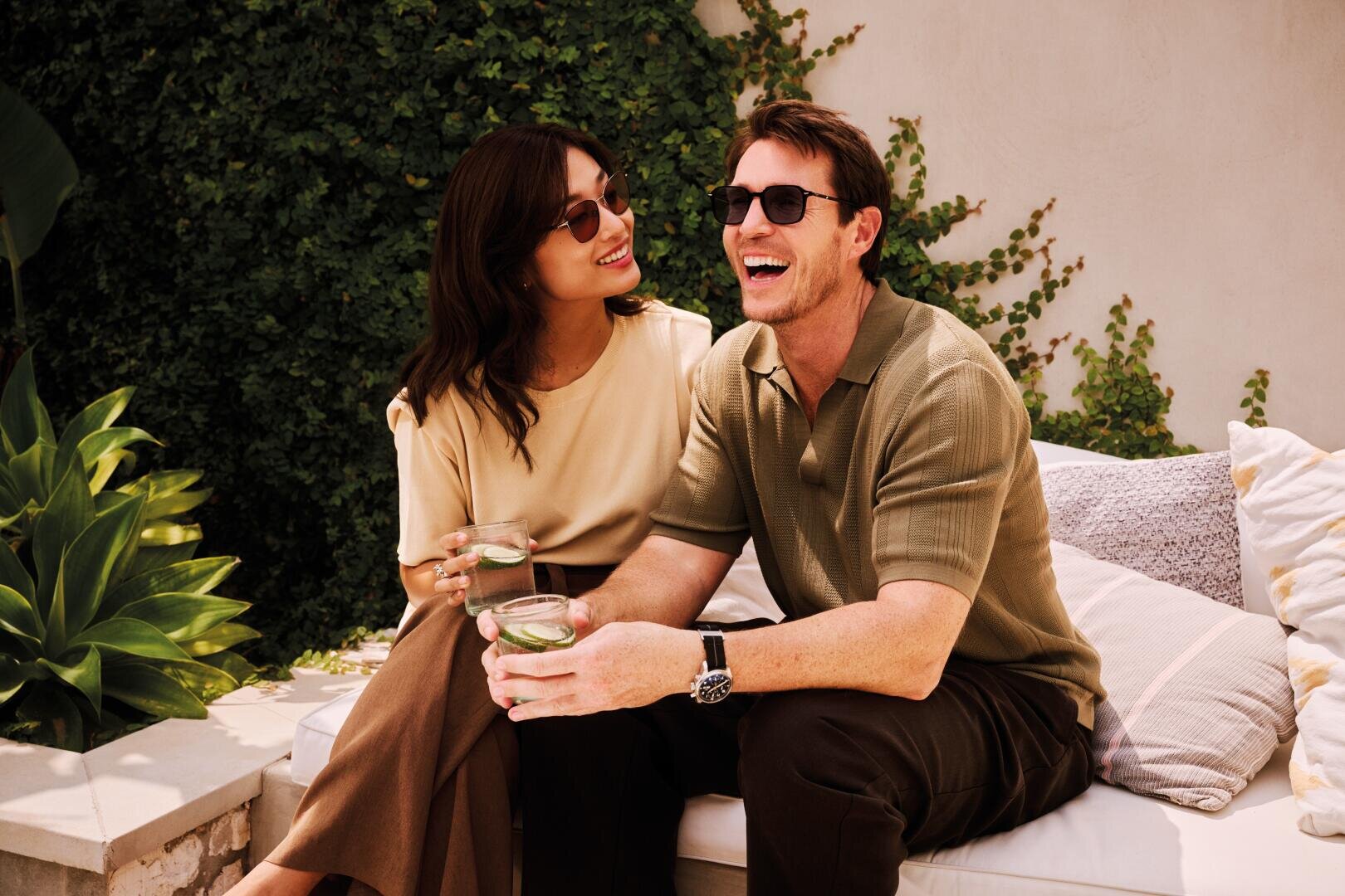 A smiling man and a woman in sunglasses sit on a couch outside, surrounded by greenery, holding drinks in their hands and enjoying a relaxing moment in the sun.
