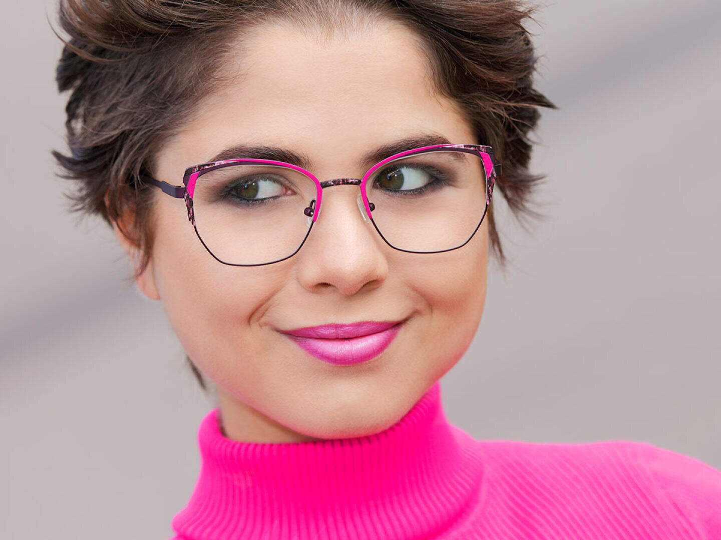 Eine Person mit kurzen braunen Haaren trägt eine große, elegante Brille und einen hellrosa Rollkragenpullover und lächelt leicht. Ihr Lippenstift passt zur Farbe ihres Pullovers.