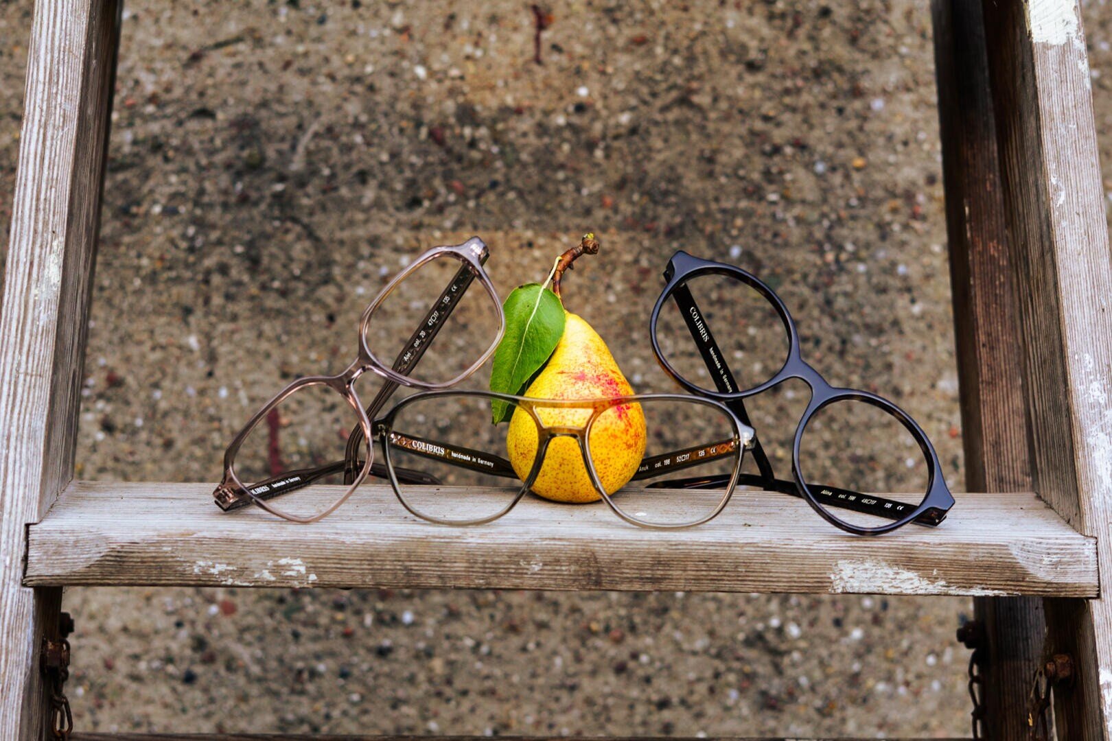 A yellow pear with a green leaf lies on a weathered wooden shelf, flanked by three pairs of glasses with different colored frames. The background is a rough, mottled gray surface.