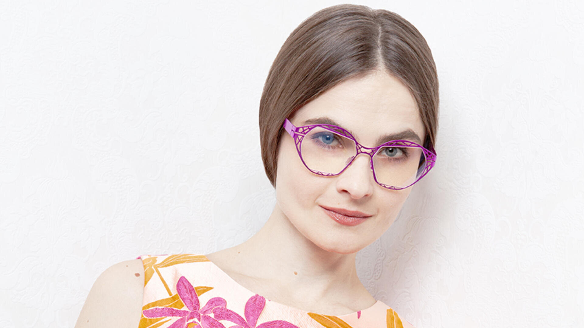 Eine Frau mit kurzen braunen Haaren, einer rosafarbenen Katzenaugenbrille und einem ärmellosen Oberteil mit buntem Blumenmuster steht vor einem hellen, strukturierten Hintergrund.