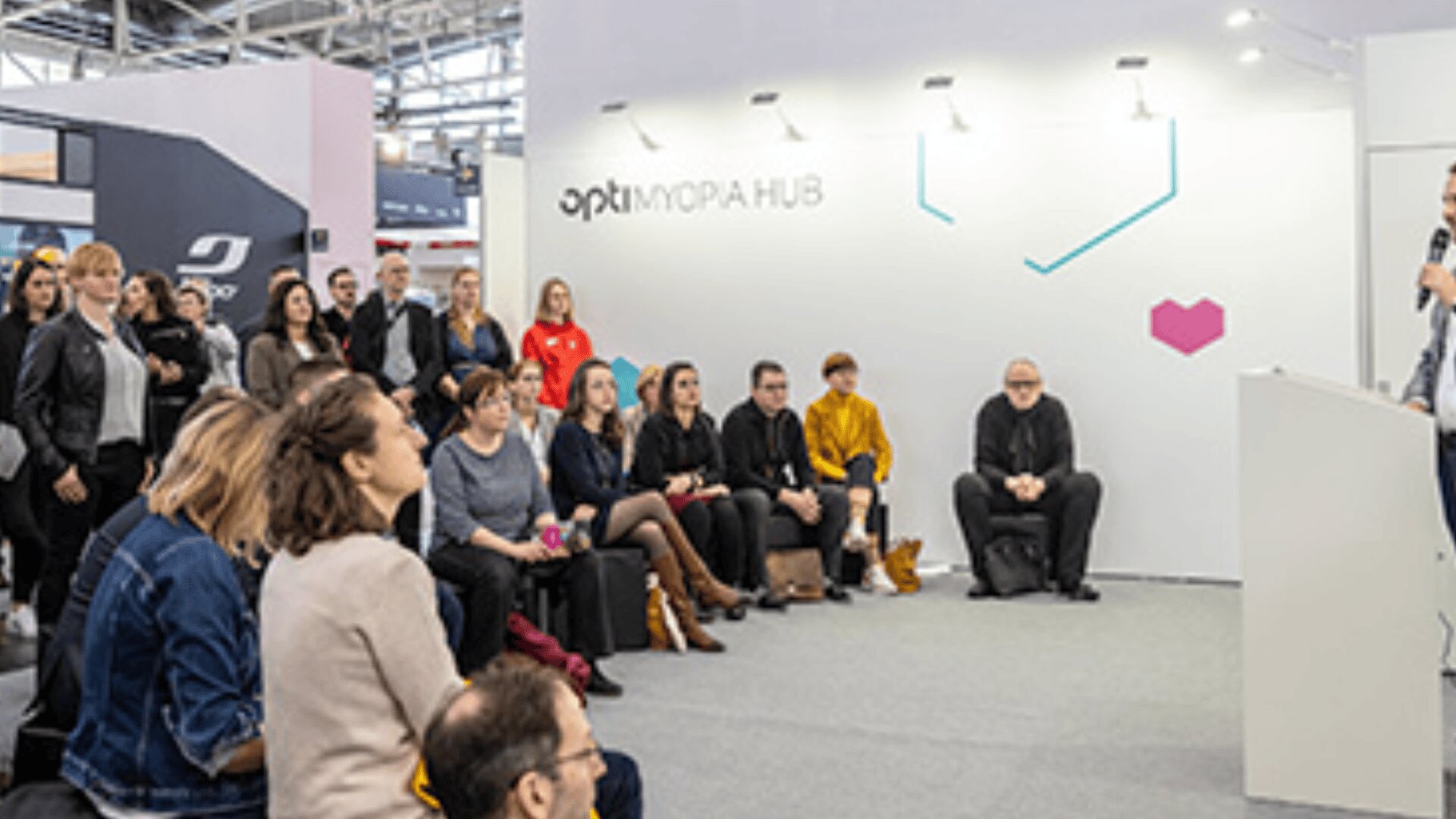 Eine Gruppe von Menschen sitzt und hört einem Redner bei einer modernen Veranstaltung oder einem Seminar in einem Gebäude aufmerksam zu. Der Rahmen besteht aus einer weißen Wand mit der Aufschrift MORPH HUB und geometrischen Mustern.