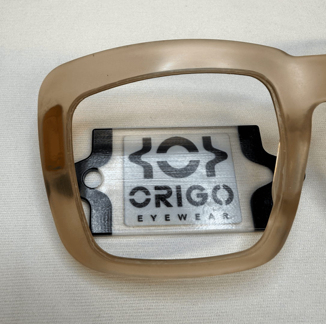 A beige frame held over a card with the logo and the text ORIGO EYEWEAR on a white surface. The lens enlarges and emphasizes the text and logo underneath.
