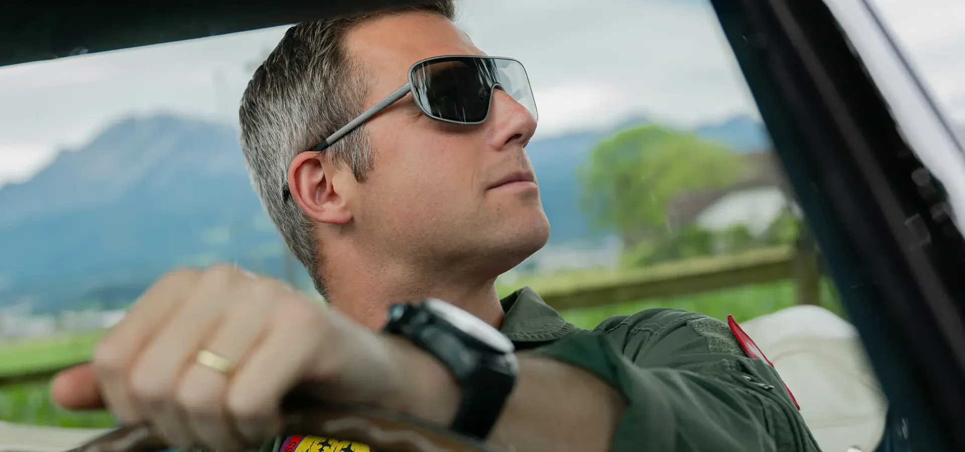 A man with sunglasses and a watch is driving a car and looking out of the window. He is wearing a green jacket and has short hair. Blurred green areas and distant mountains can be seen in the background.