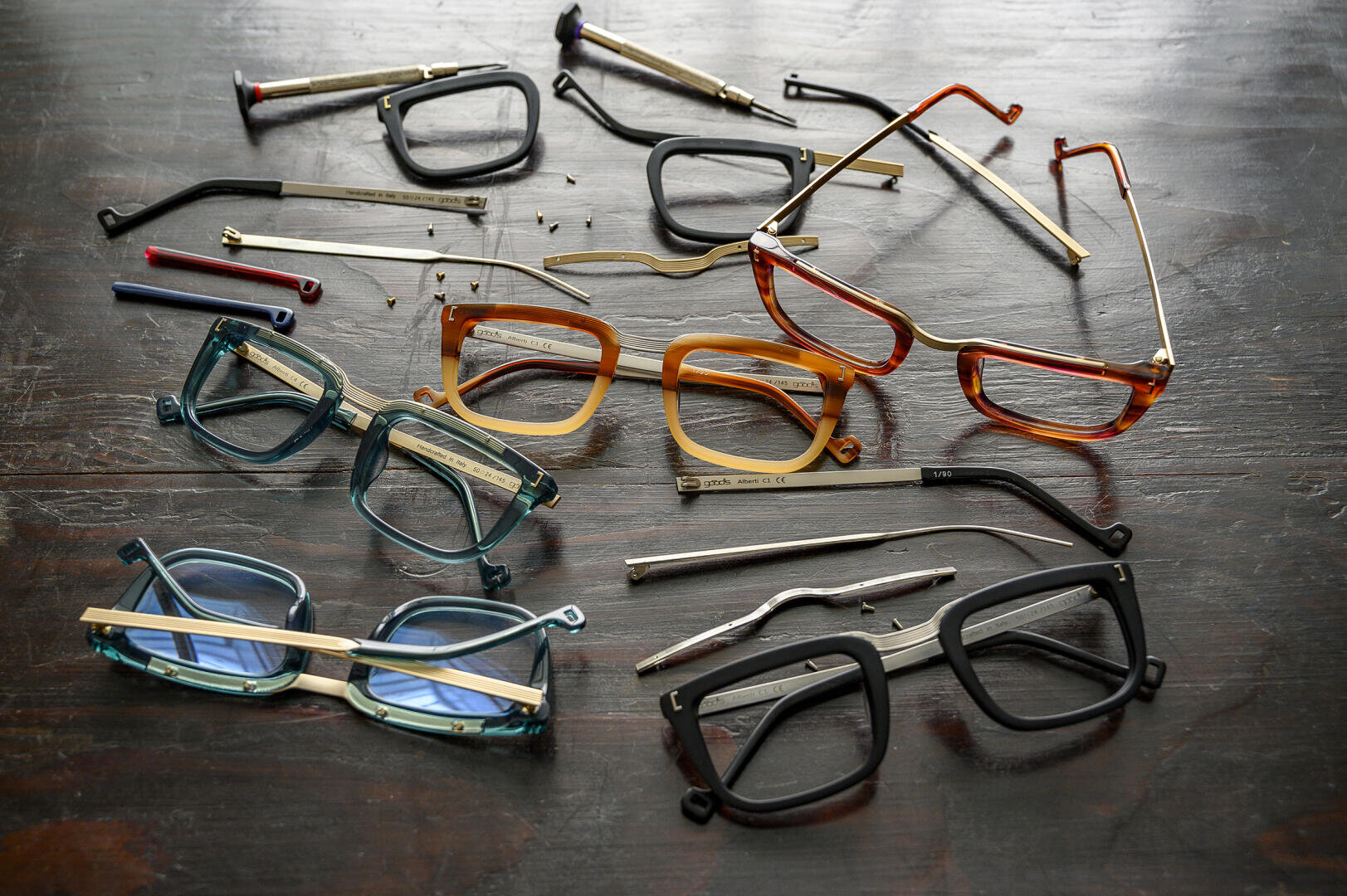 Several frames and temples in different colors and styles are disassembled and spread out on a dark wooden surface, with small screws scattered between the pieces.