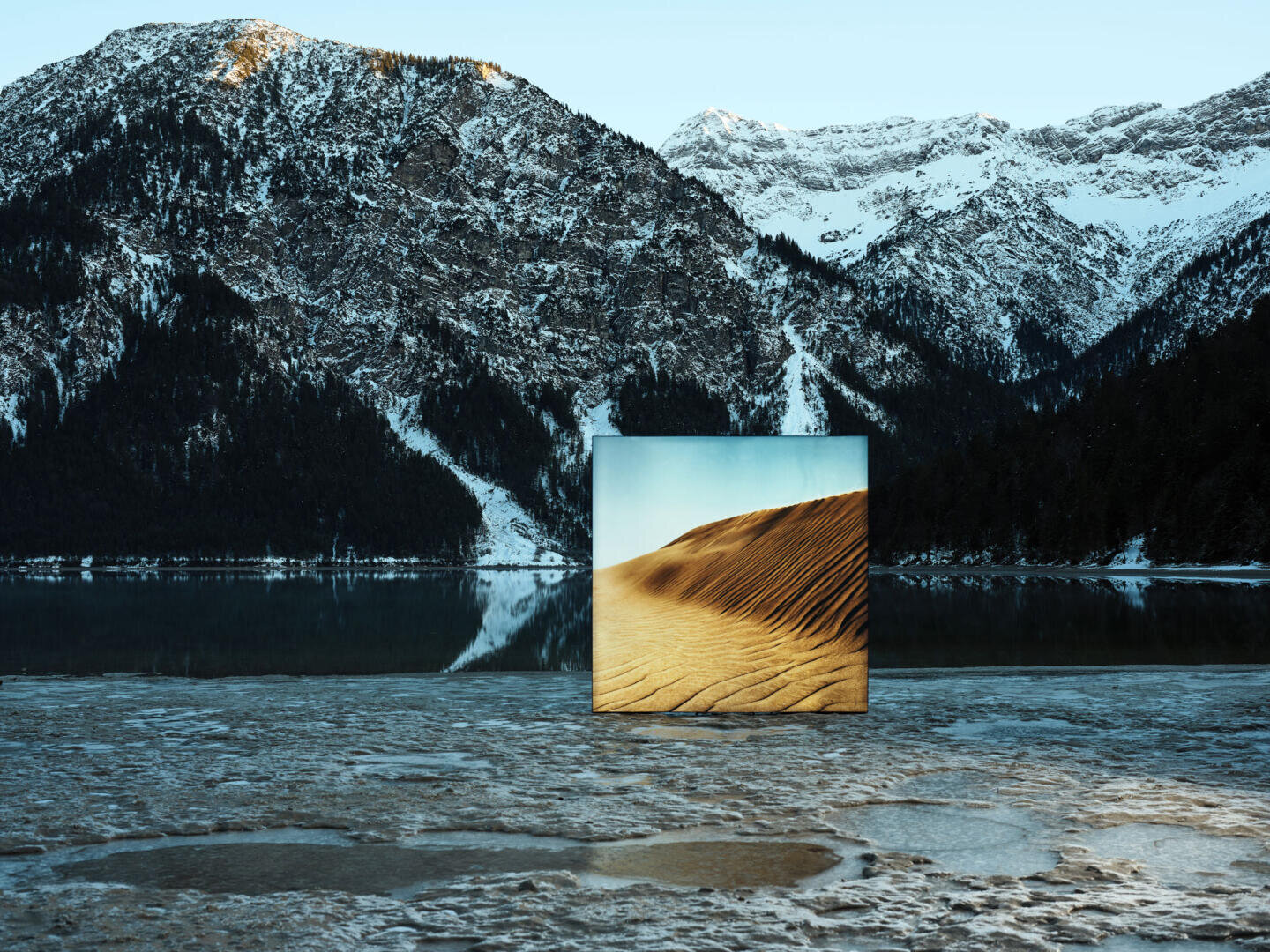 Ein quadratischer Spiegel steht auf einem verschneiten, zugefrorenen See, der von Bergen umgeben ist, und reflektiert das Bild einer sonnigen Wüstensanddüne, das in starkem Kontrast zur winterlichen Landschaft steht.