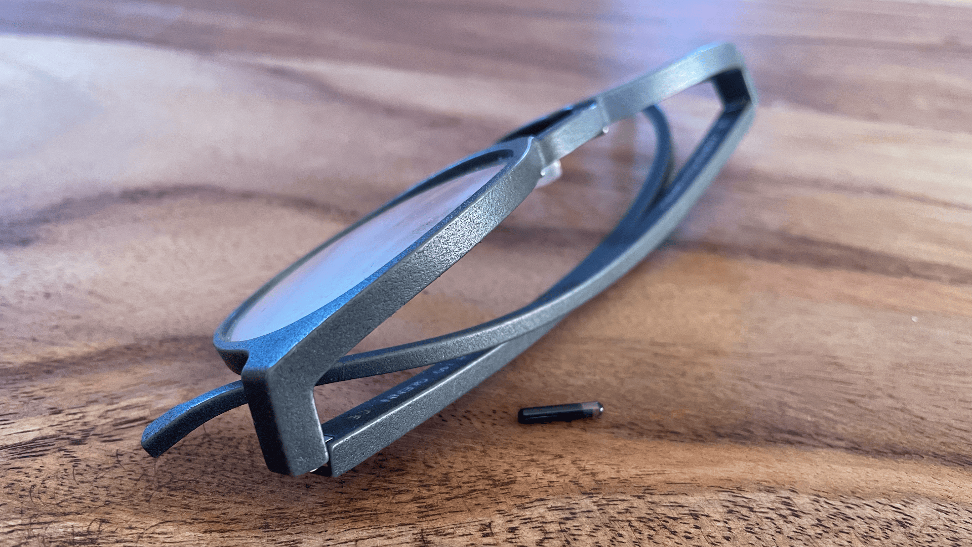 Eine Brille mit schwarzem Rahmen liegt auf einer Holzoberfläche. Einer der Bügel ist abgenommen und neben dem Rahmen liegt eine kleine Schraube.
