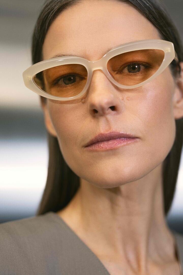 A woman with straight brown hair wears beige cat-eye sunglasses with brown lenses and a neutral expression, dressed in a gray top, against a softly blurred background.