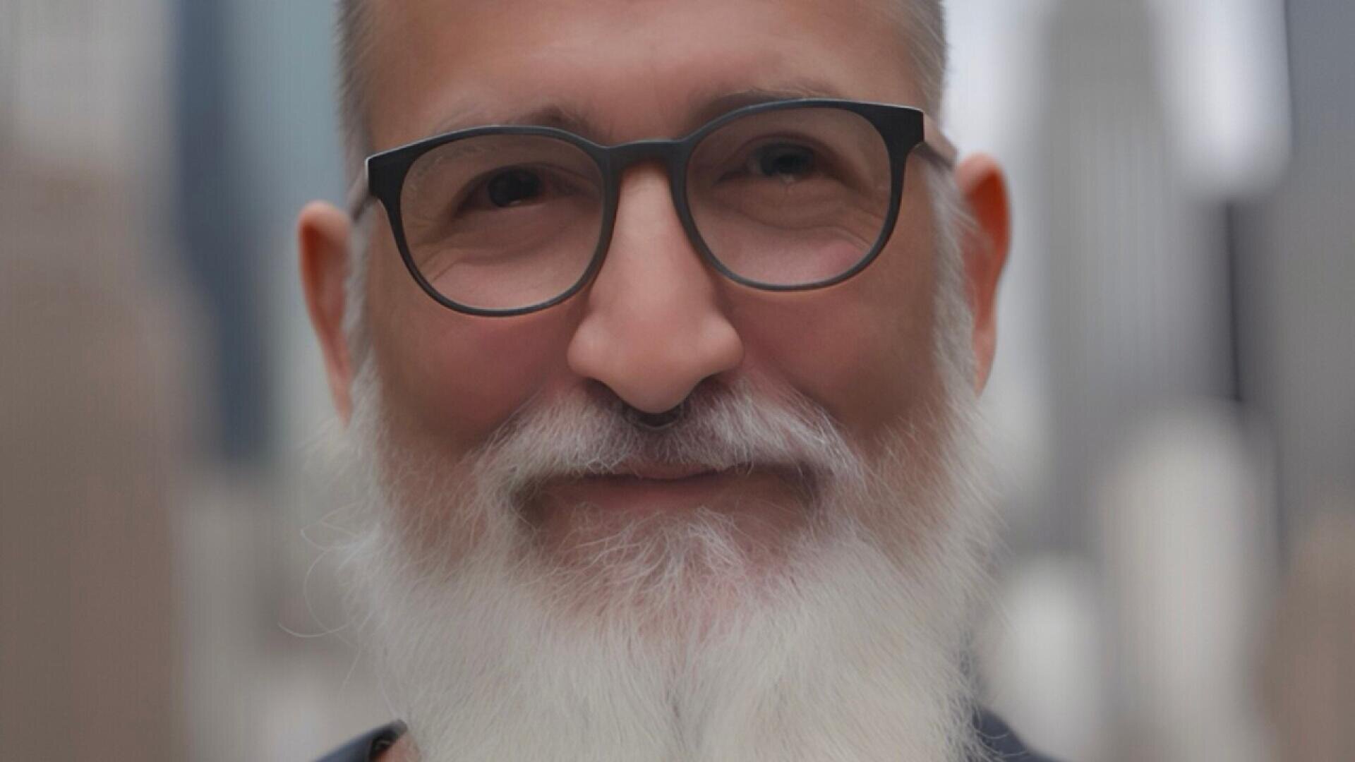A smiling older man with a full white beard and mustache, wearing dark-rimmed glasses, standing outdoors with blurred city buildings in the background.