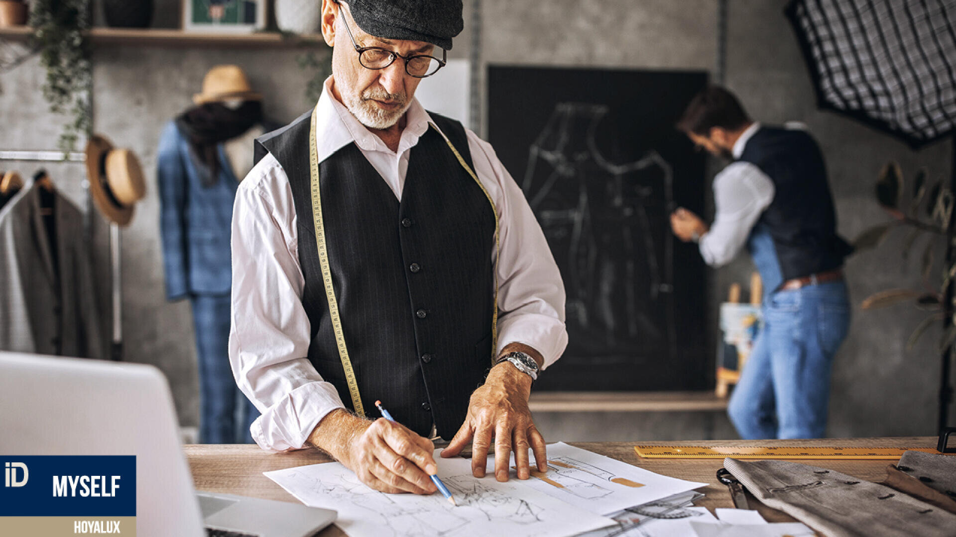 Ein älterer Mann in Weste, Brille und Schiebermütze skizziert an einem Arbeitstisch Kleidungsentwürfe, ein Maßband um den Hals. Im Hintergrund einer Schneiderwerkstatt arbeitet ein zweiter Mann an einer Tafel.