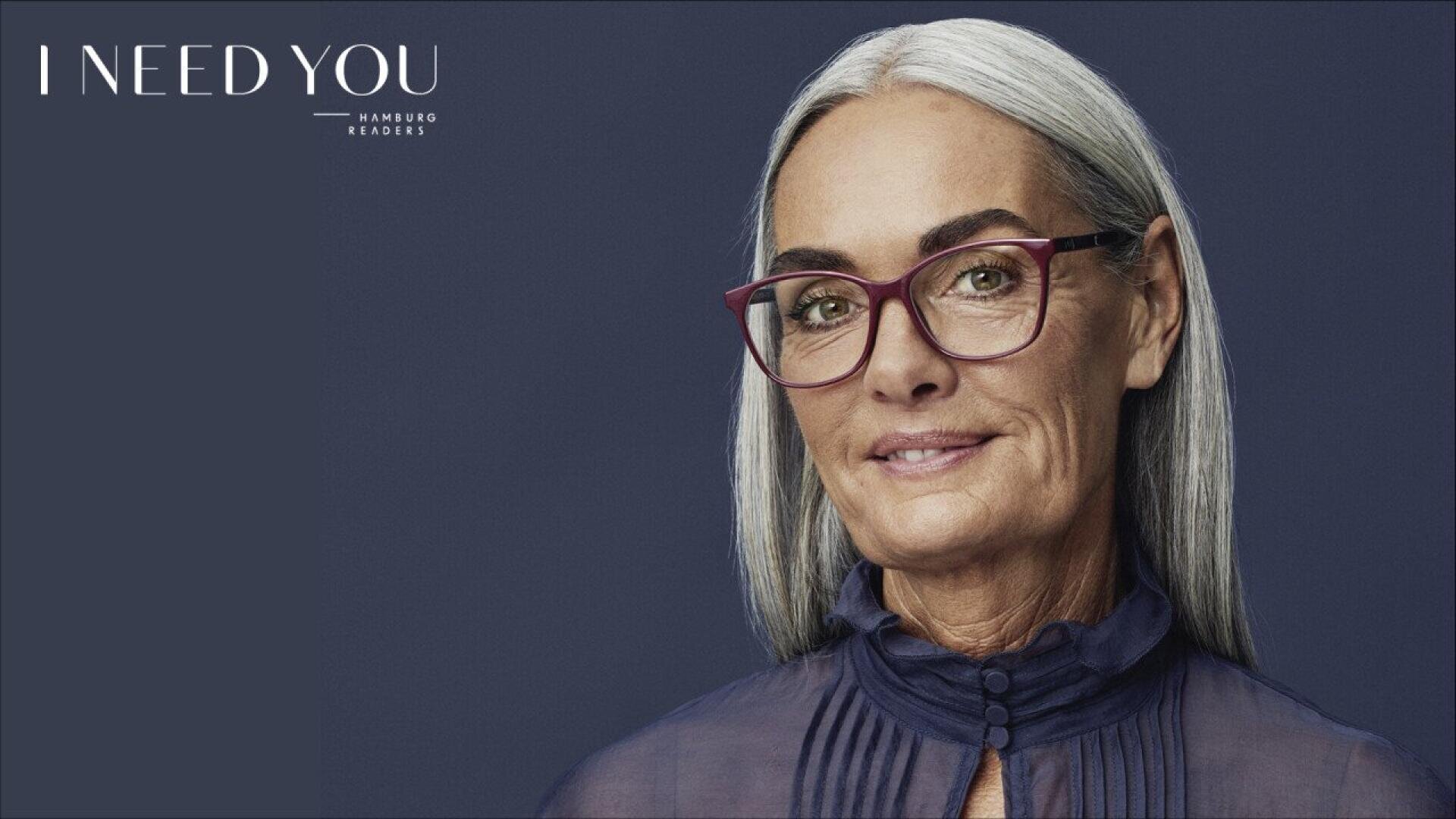 An older woman with long gray hair and glasses smiles softly against a dark background. She is wearing a sheer, high-neck blouse. Text in the top left corner reads I NEED YOU Hamburg Readers.