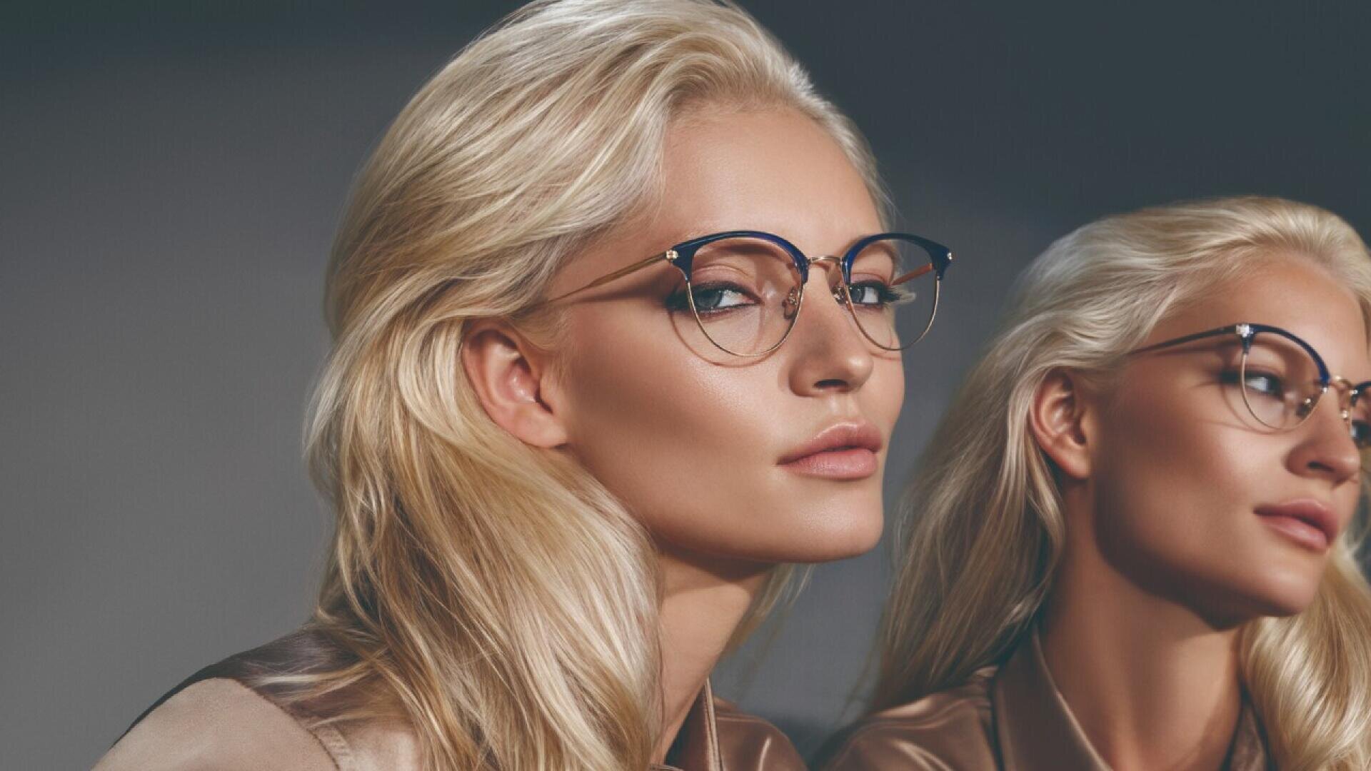 A blonde woman with long hair and glasses looks confidently at the camera, with her mirror reflection beside her against a neutral background. Both wear tan satin tops and appear calm and composed.