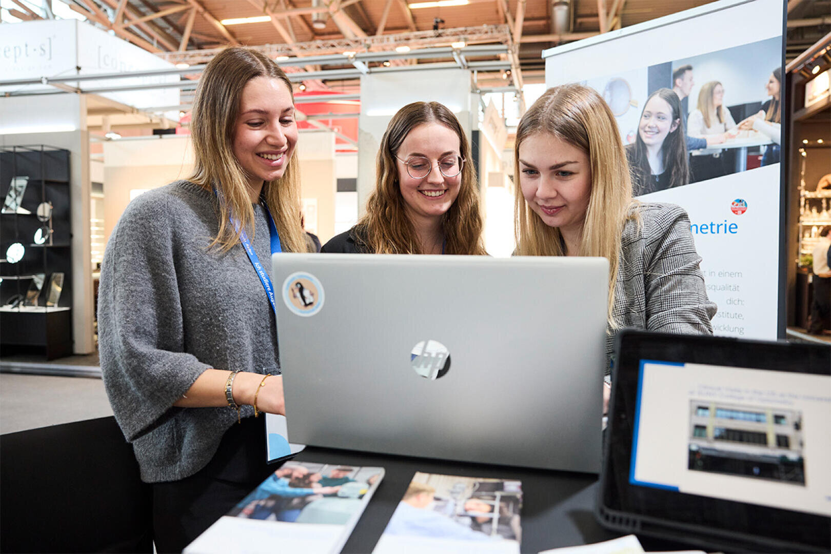 Drei junge Frauen stehen zusammen, lächeln und blicken auf einen Laptop auf einem Tisch, bei dem es sich anscheinend um eine berufliche Veranstaltung oder Konferenz handelt. Auf dem Tisch vor ihnen sind auch Flyer und ein Tablet zu sehen.
