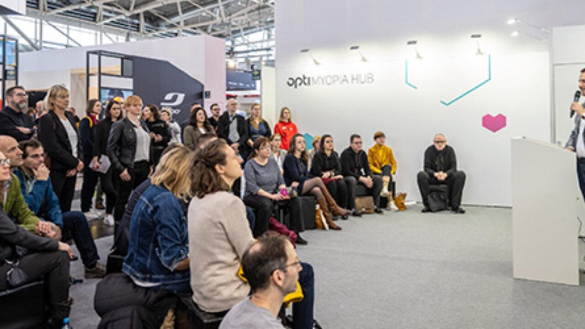 A group of people sit and stand attentively, listening to a speaker at a podium in a modern, well-lit exhibition space with a sign that reads OPTIMORA HUB on the wall.