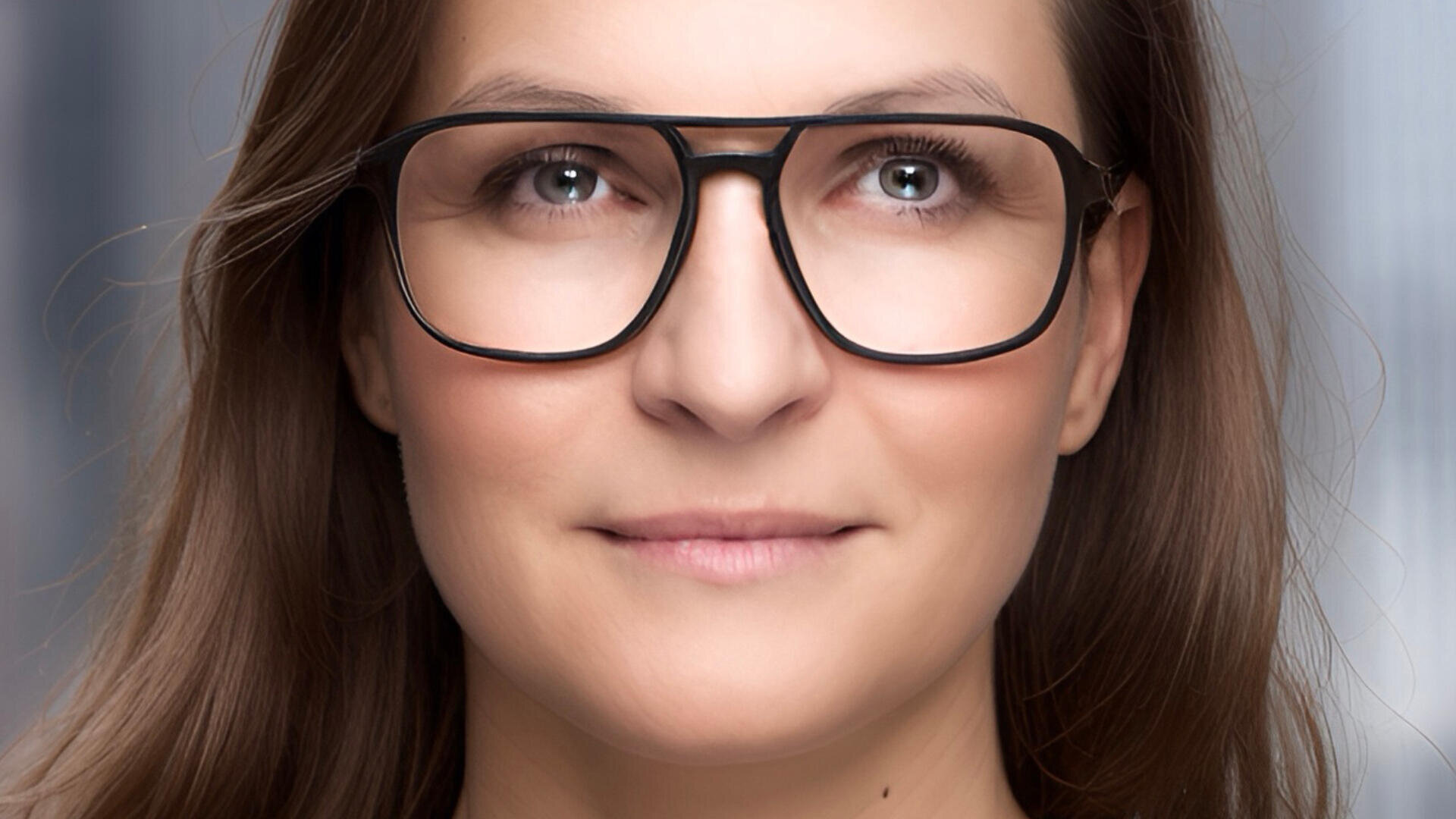 Eine Frau mit langen braunen Haaren und heller Haut trägt eine große Brille mit schwarzem Rand und lächelt leicht. Der Hintergrund ist sanft in Grau- und Blautönen verschwommen.
